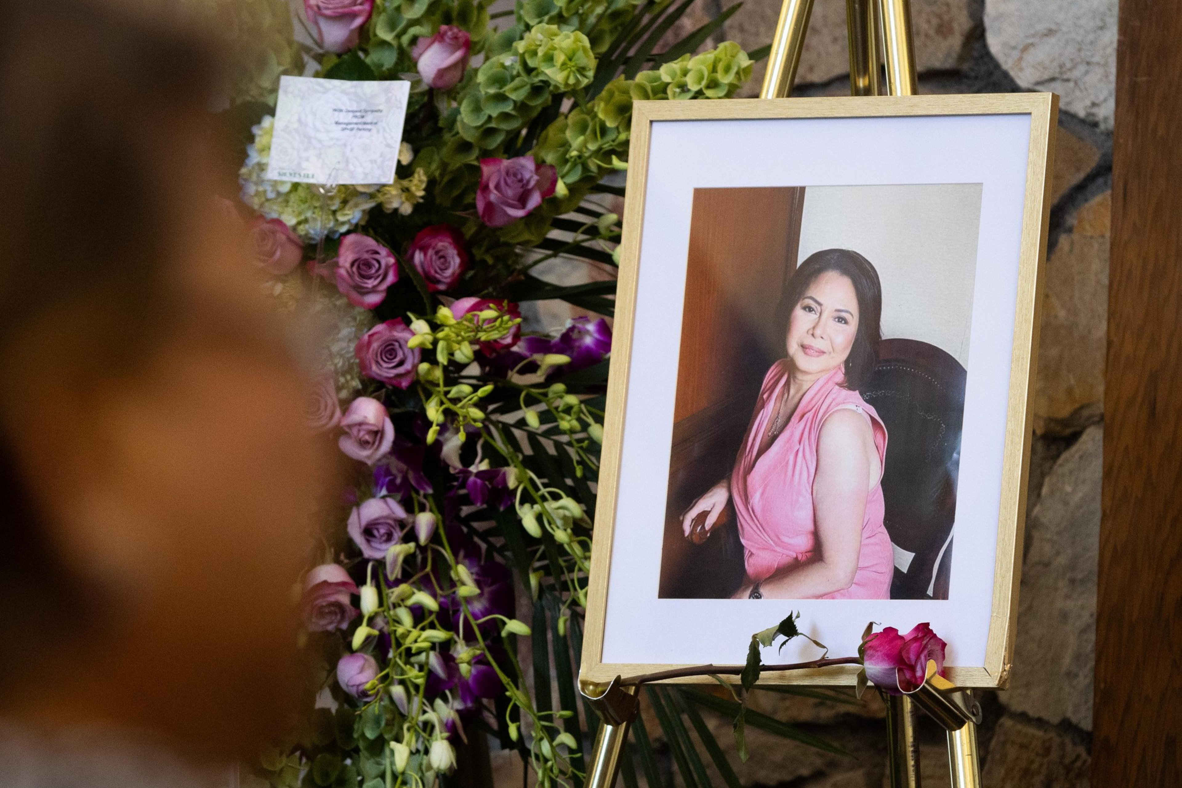 A framed photo of a woman in a pink dress is displayed on an easel next to a bouquet of purple and green flowers. A person is blurred in the foreground.