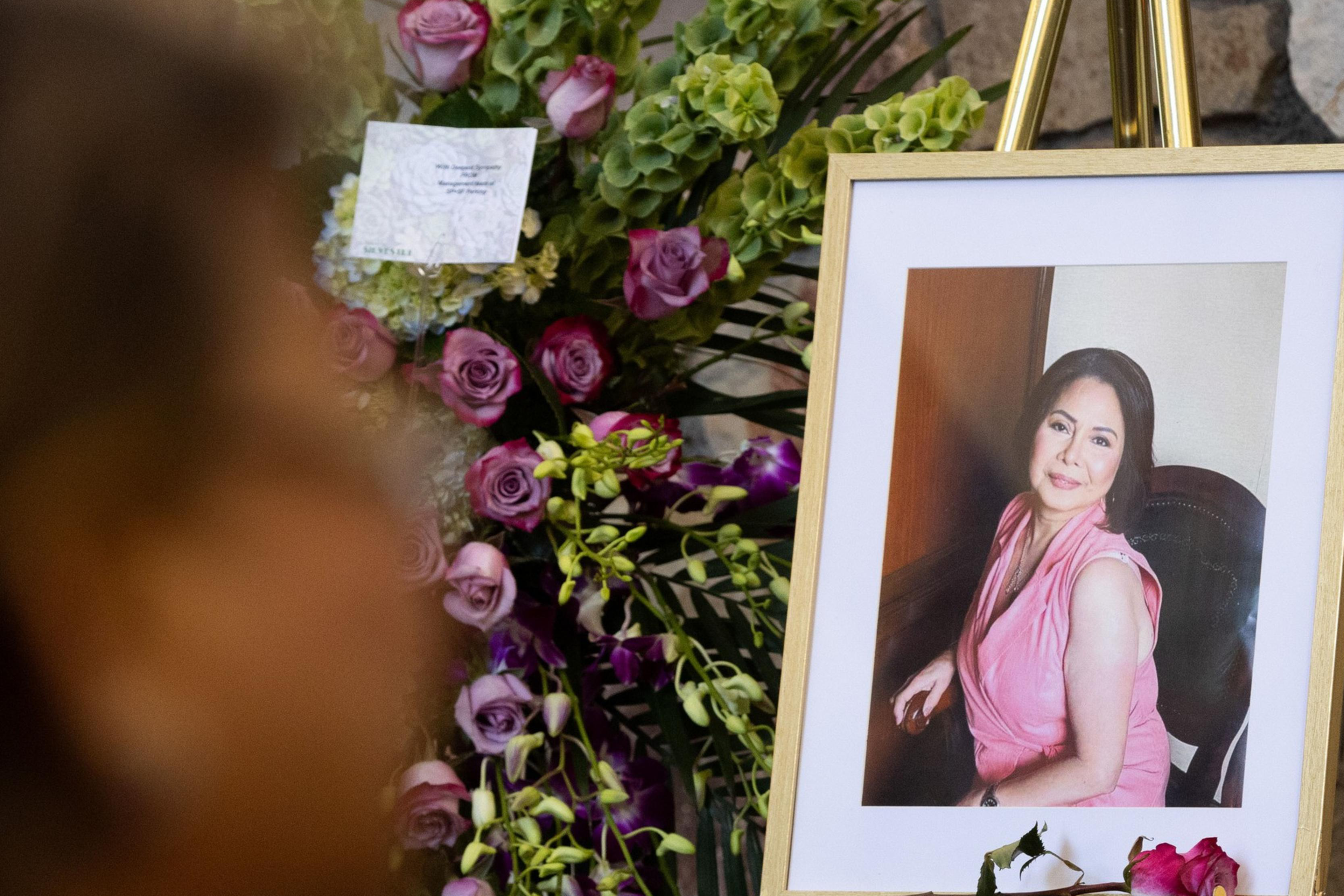 A framed photo of a woman in a pink dress is displayed on an easel next to a bouquet of purple and green flowers. A person is blurred in the foreground.