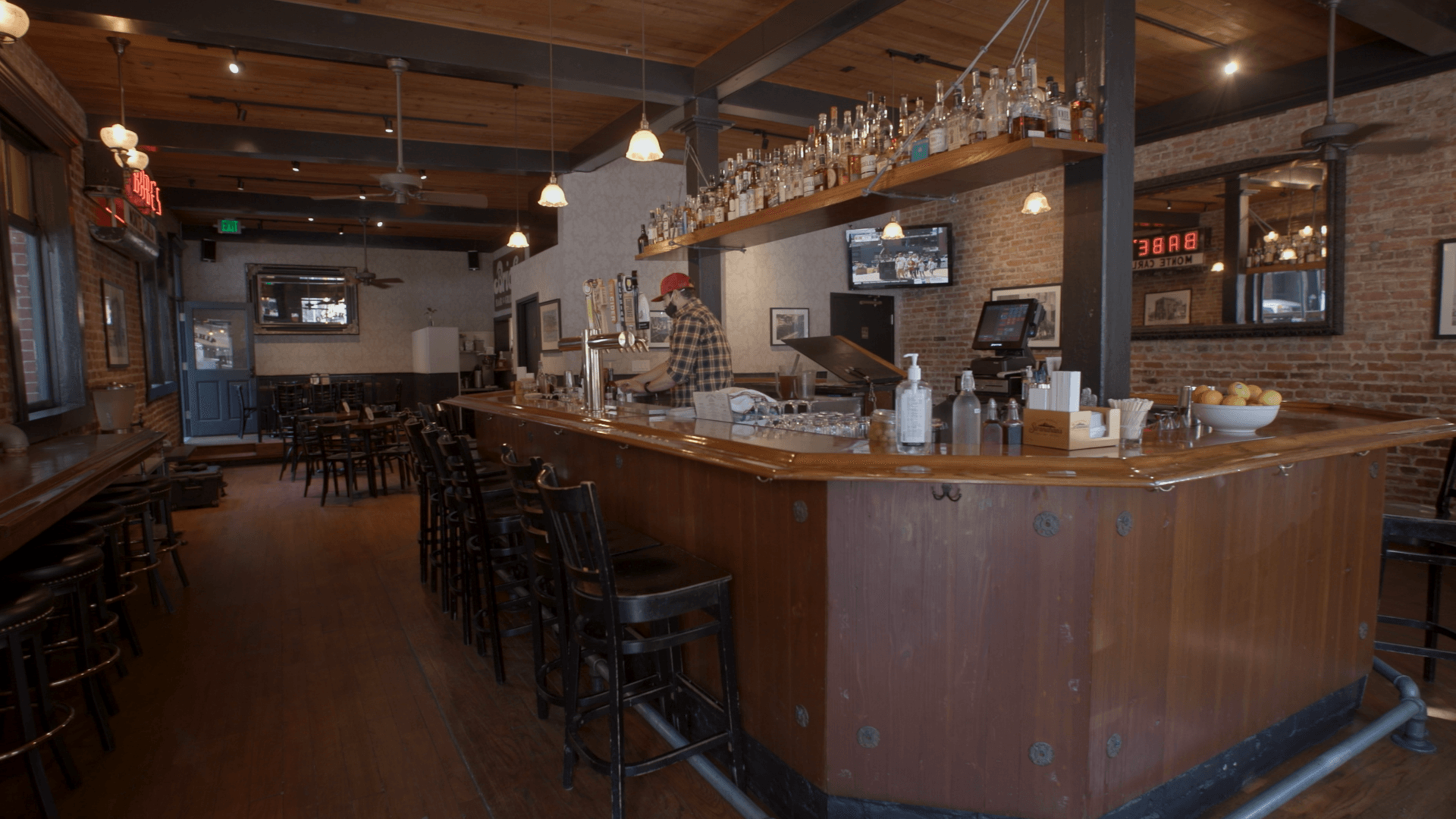 A wooden bar with stools surrounds a bartender pouring drinks, with shelves of bottles overhead, brick walls, dim lights, and tables in the background.