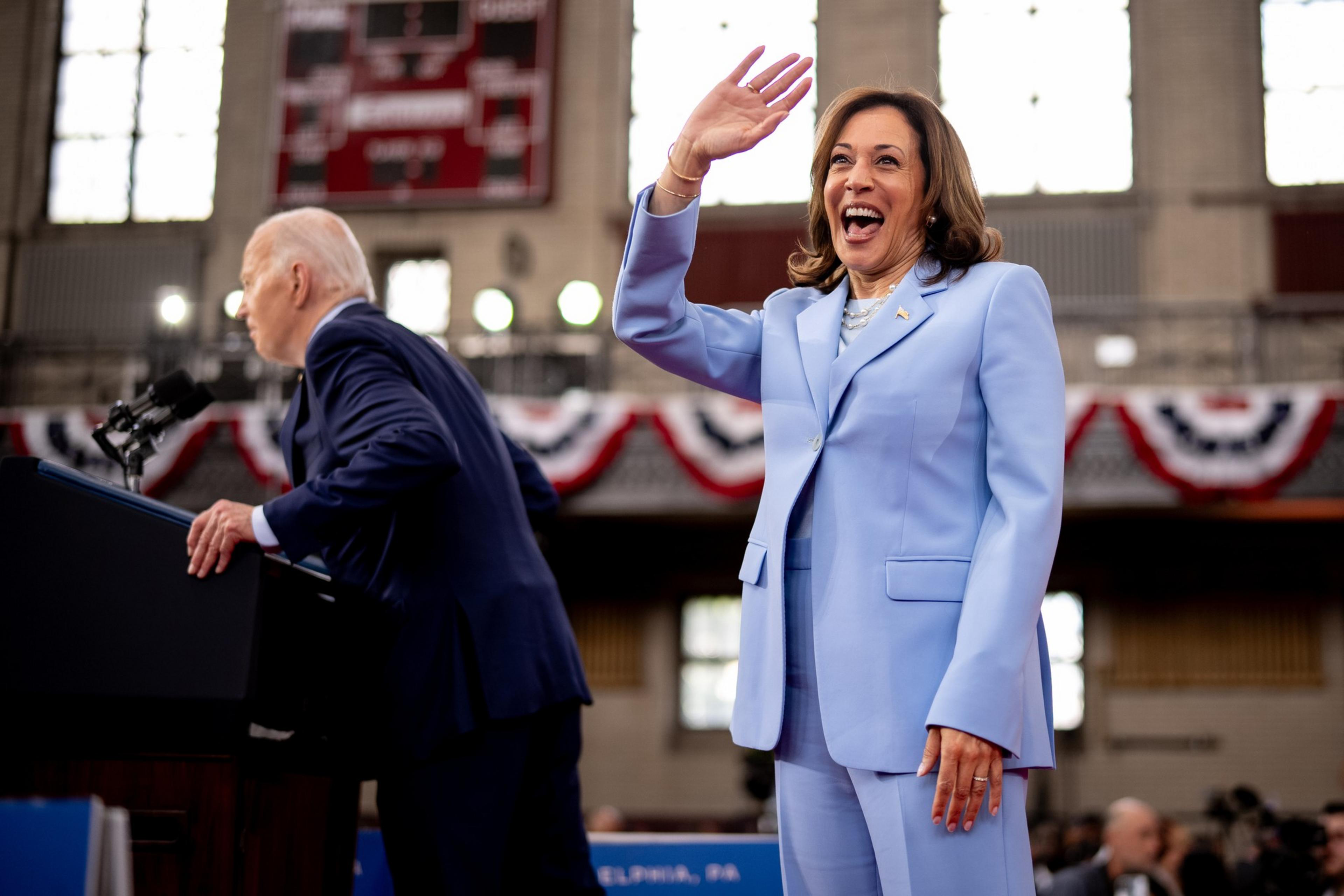 A woman in a light blue suit smiles and waves, standing behind a man in a dark blue suit who is leaning over a podium. The background has flags and a scoreboard.