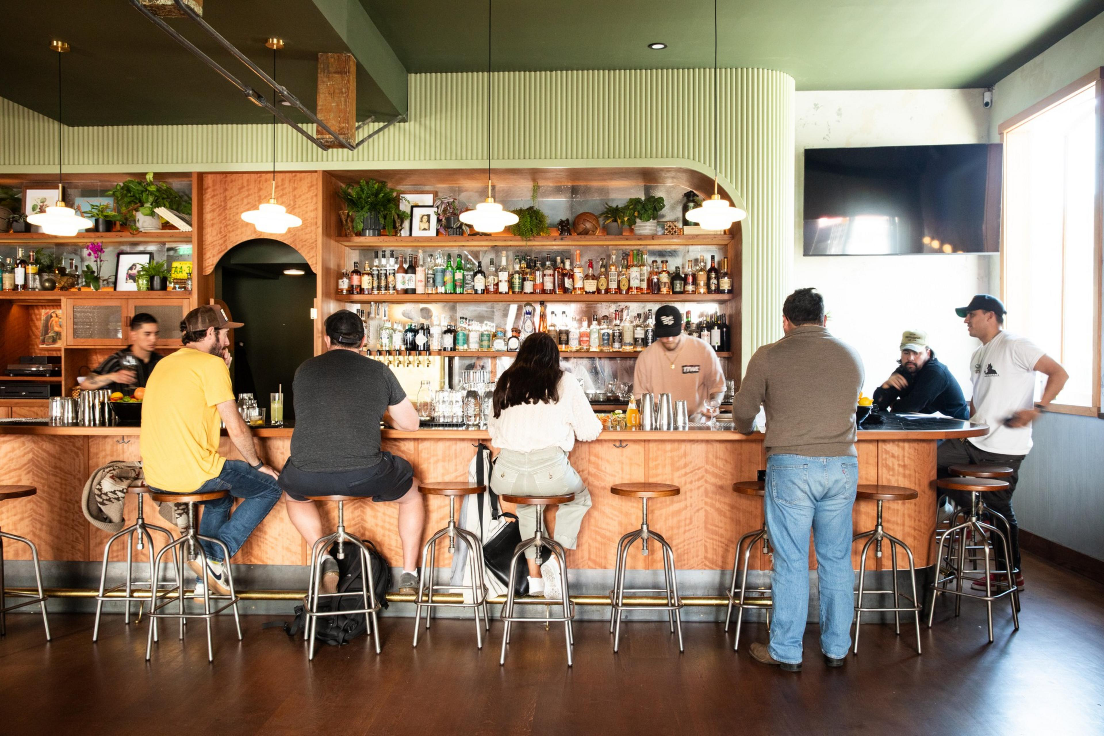 Three people sit at a bar counter surrounded by numerous bottles and plants, while two bartenders prepare drinks. The ambiance is cozy with warm lighting.