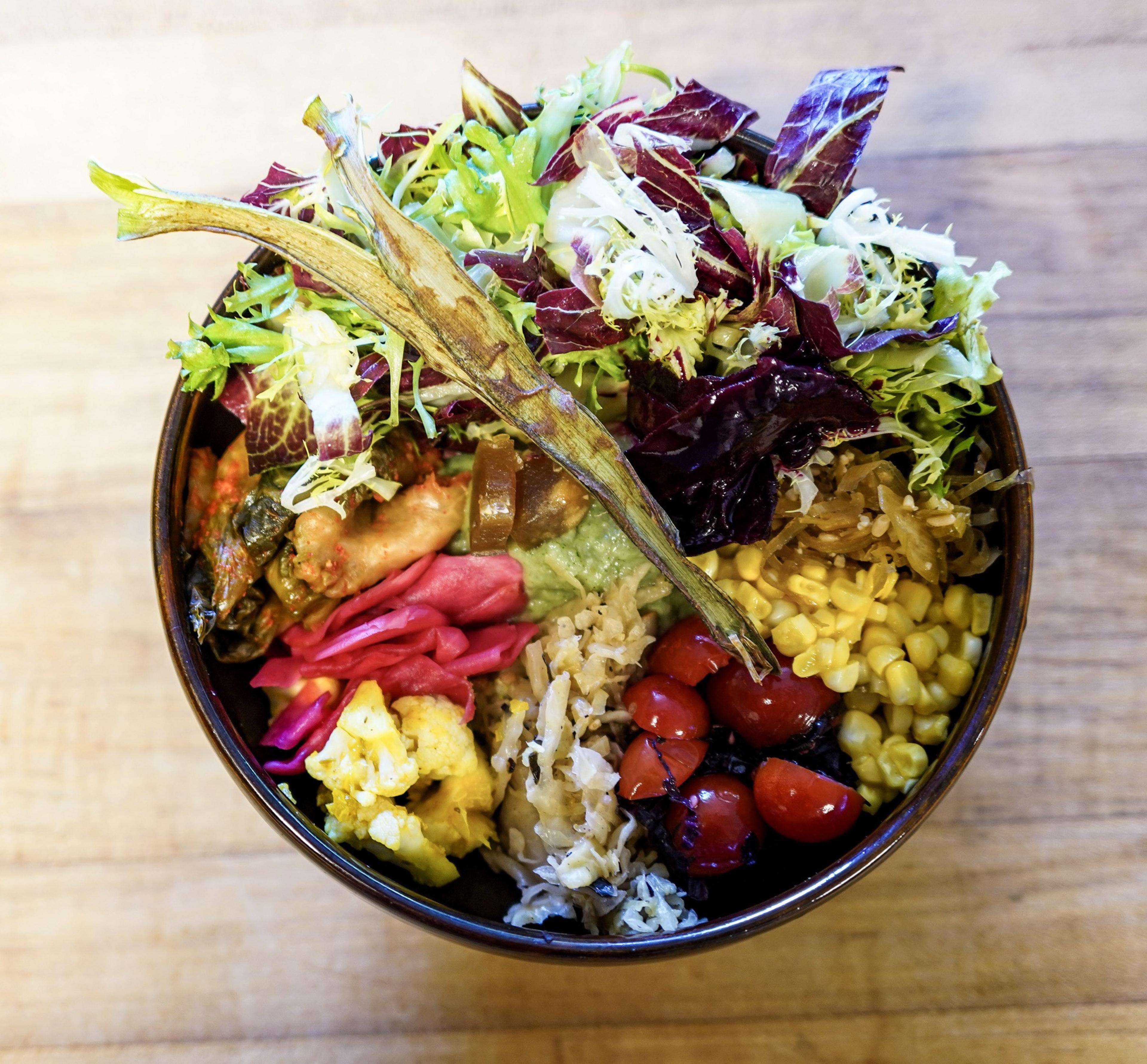 A bowl contains mixed greens, corn, cherry tomatoes, pickled vegetables, cauliflower, cooked rice, and a dried okra pod on top.