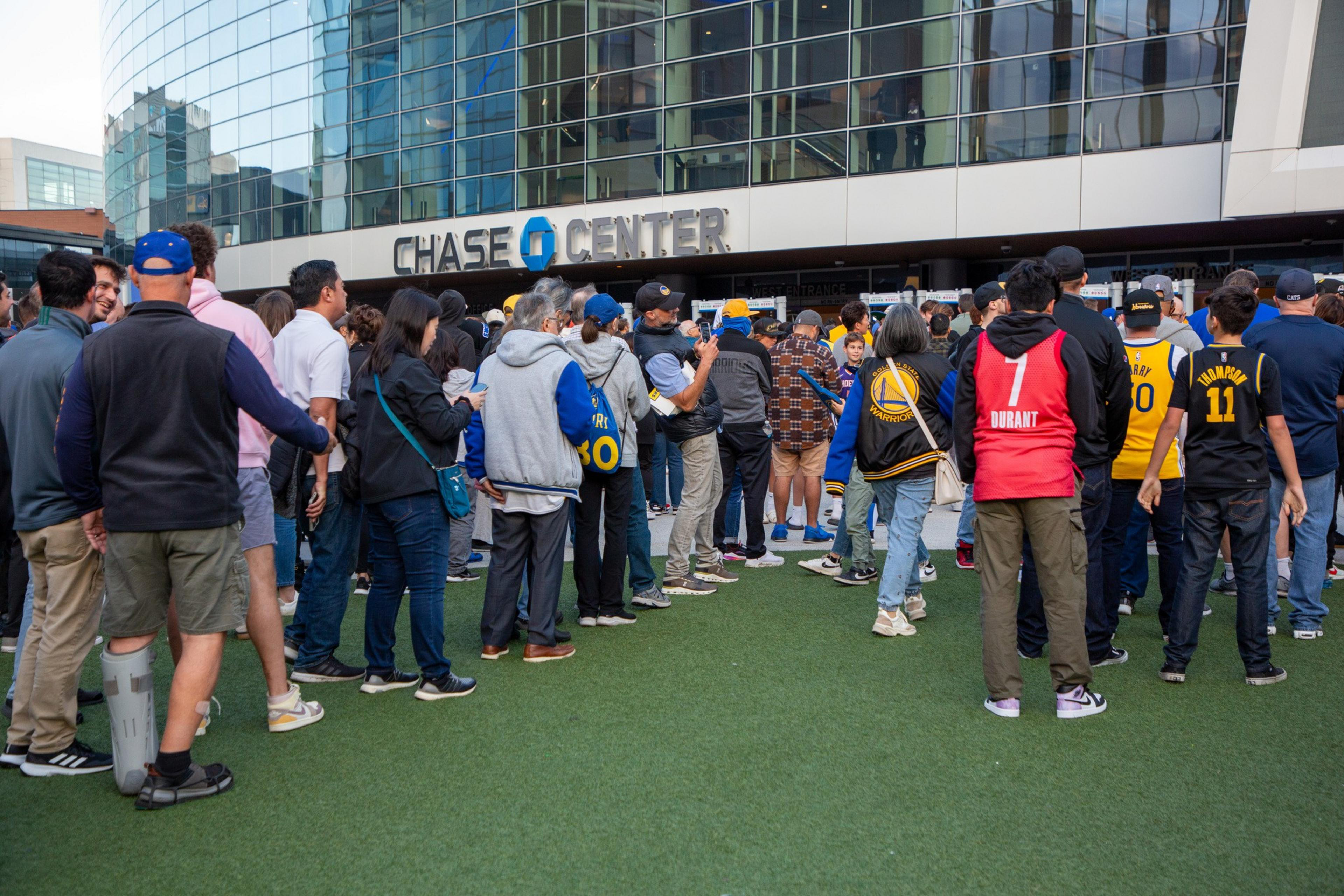 A crowd of people, many wearing basketball jerseys, is gathered outside the Chase Center, a modern glass building. The atmosphere is lively and social.