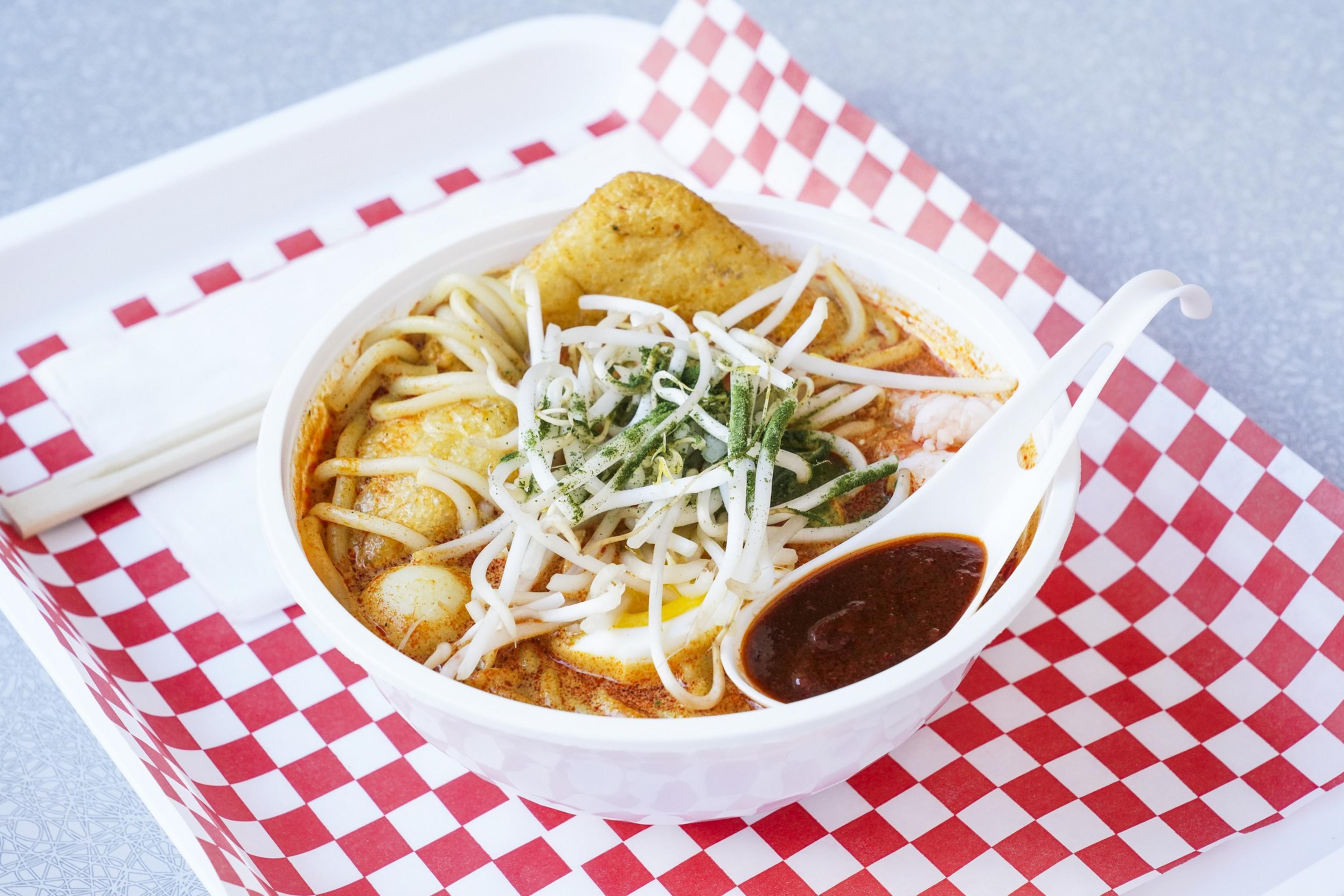 A bowl of noodle soup topped with bean sprouts, herbs, and a slice of fried tofu. A spoon holds a dark sauce, resting on a red-checkered tray with chopsticks.