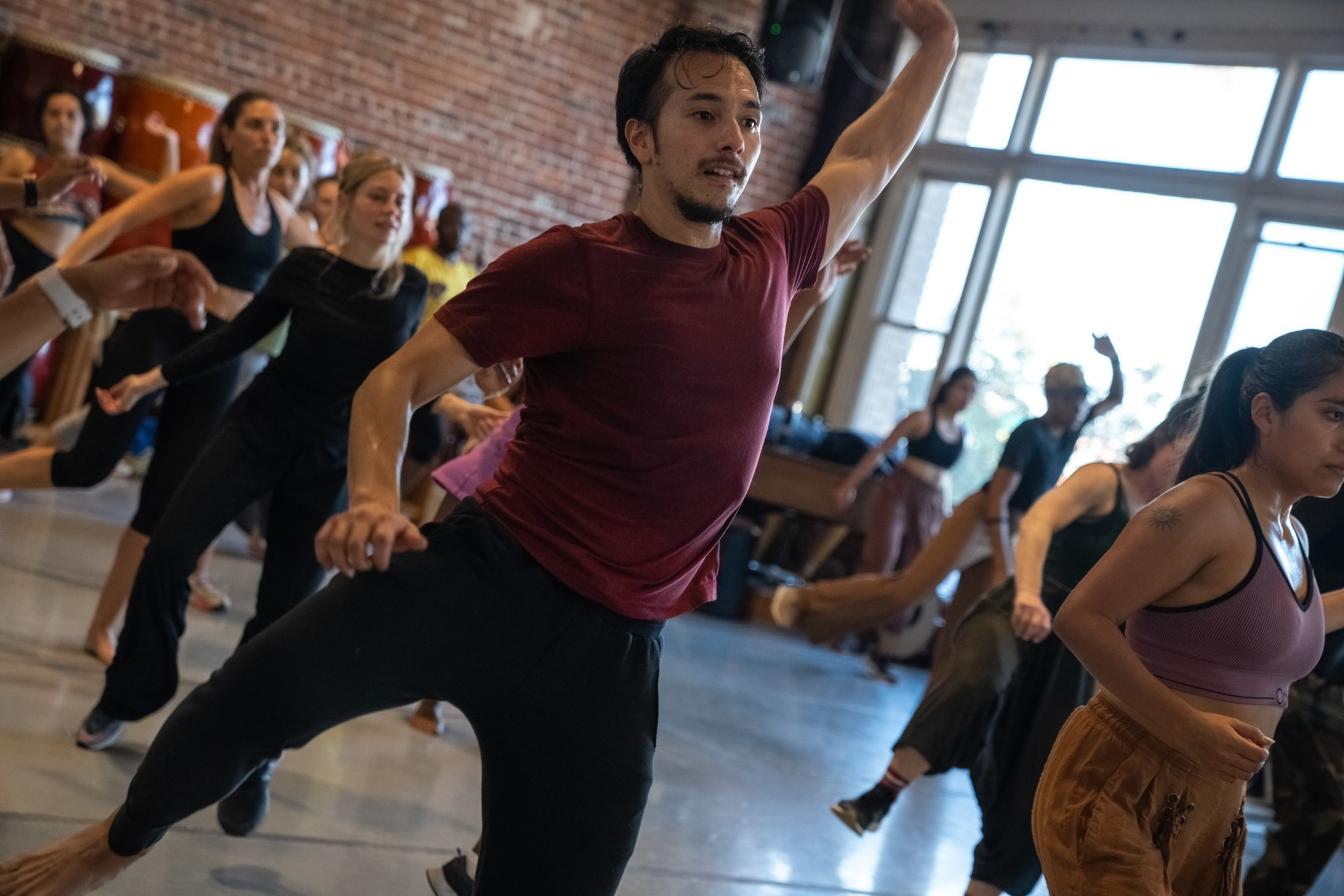 A diverse group of people are energetically dancing in a sunlit studio with large windows and a brick wall. They wear casual, comfortable clothing.