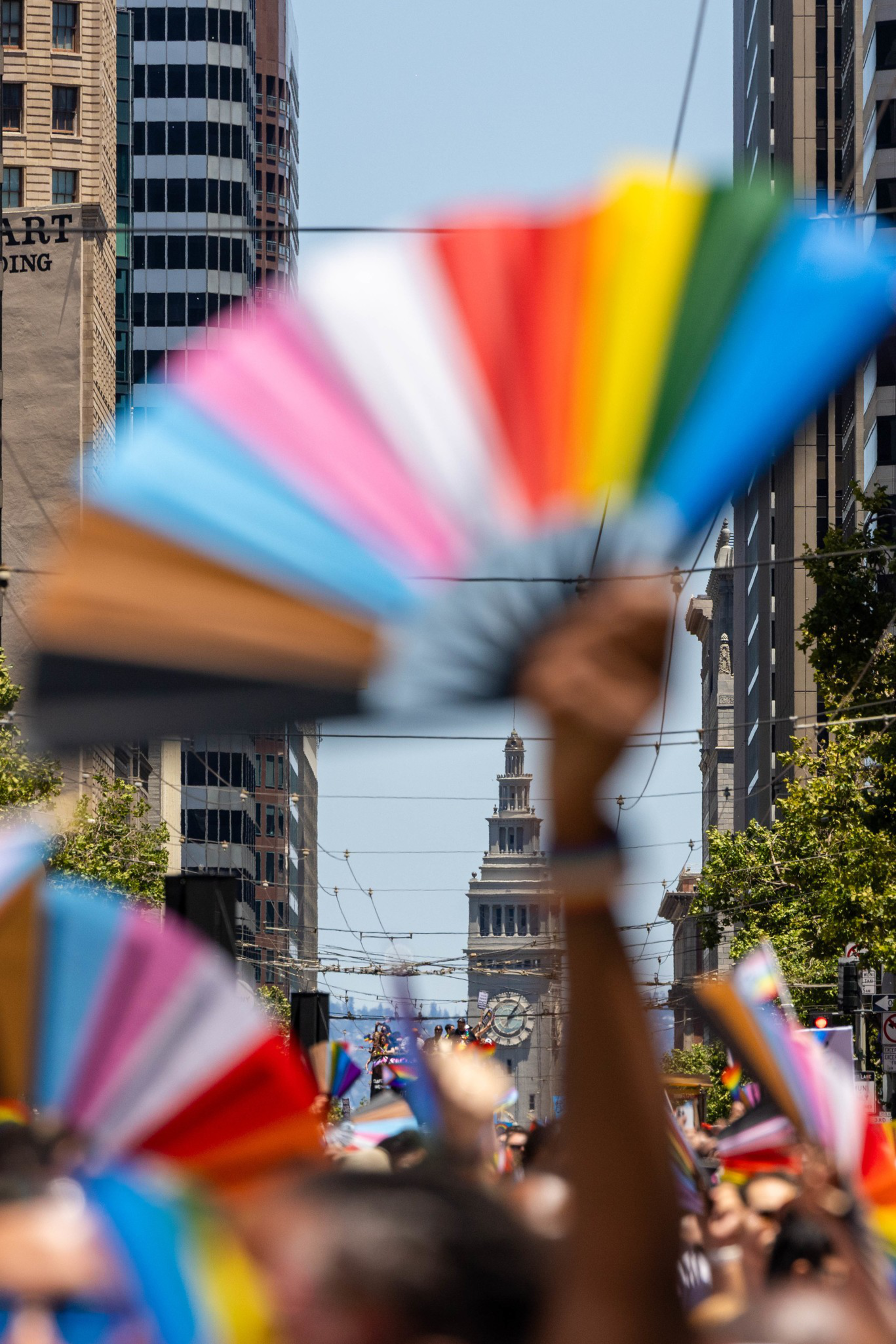 The image shows a vibrant city street scene with people holding colorful, rainbow-themed fans. The background features a prominent tower amidst tall buildings.