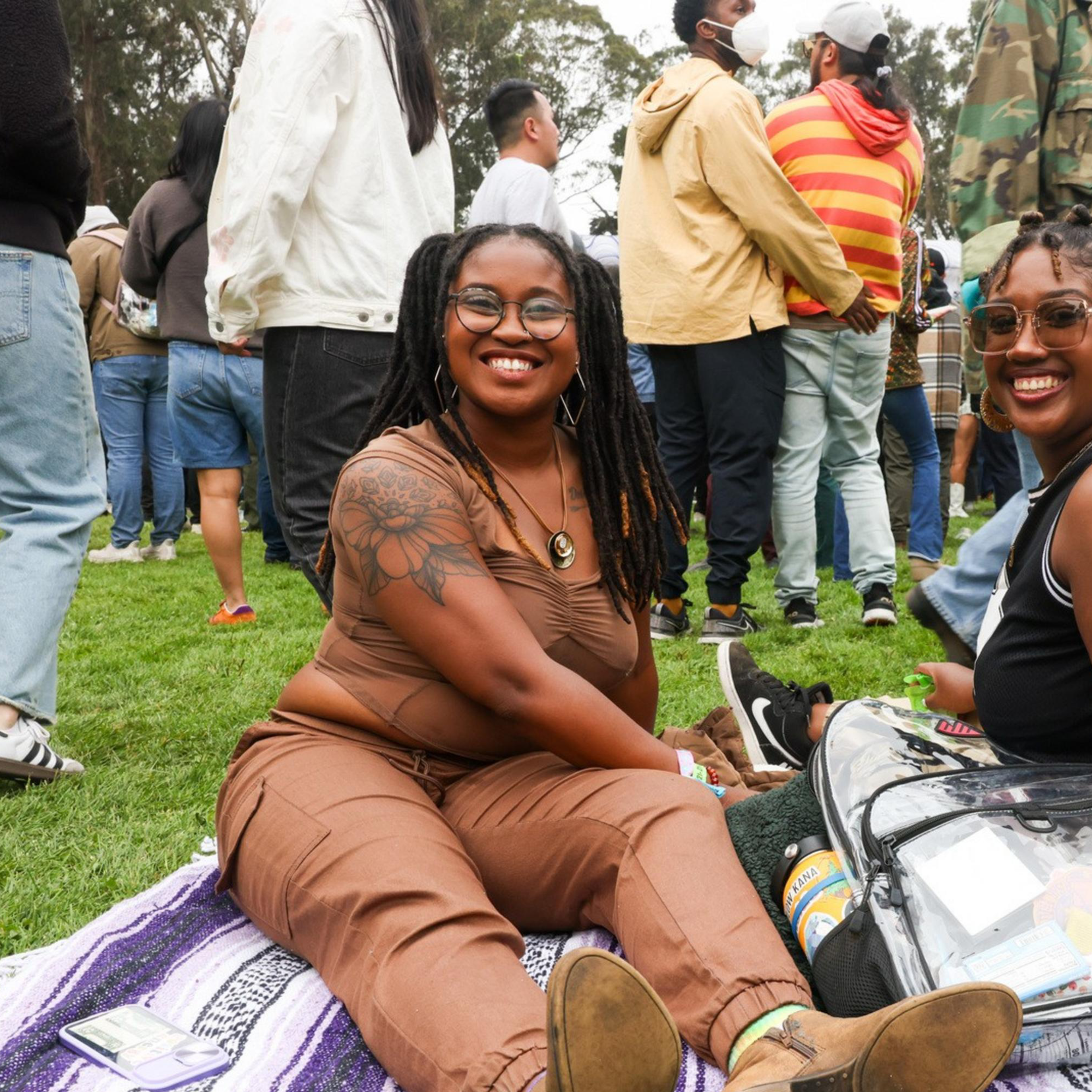 Two women sit on a blanket at an outdoor event, smiling at the camera, surrounded by a crowd of people. Both seem relaxed and happy, enjoying the atmosphere.