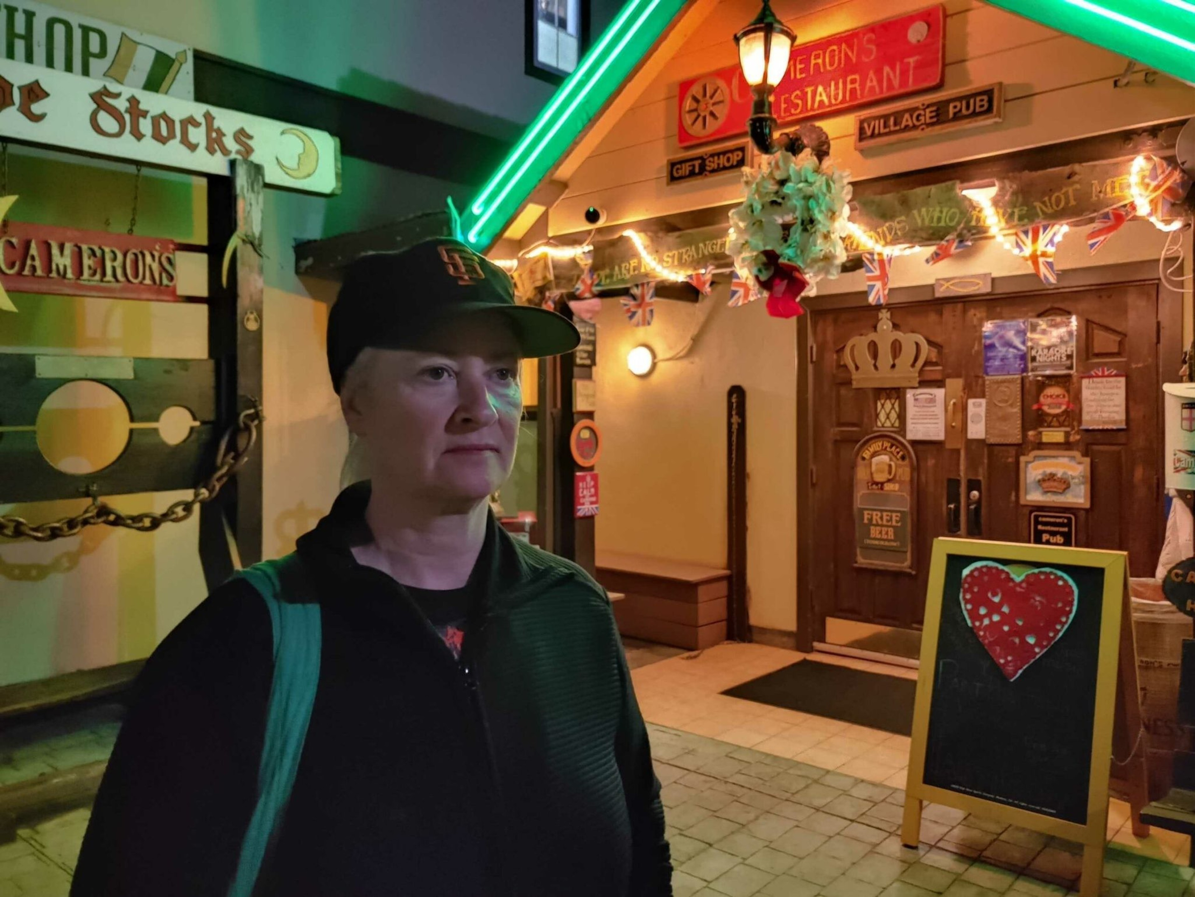 A person in a black cap and jacket stands outside a warmly lit pub decorated with hanging flowers, British flags, and a chalkboard with a red heart.