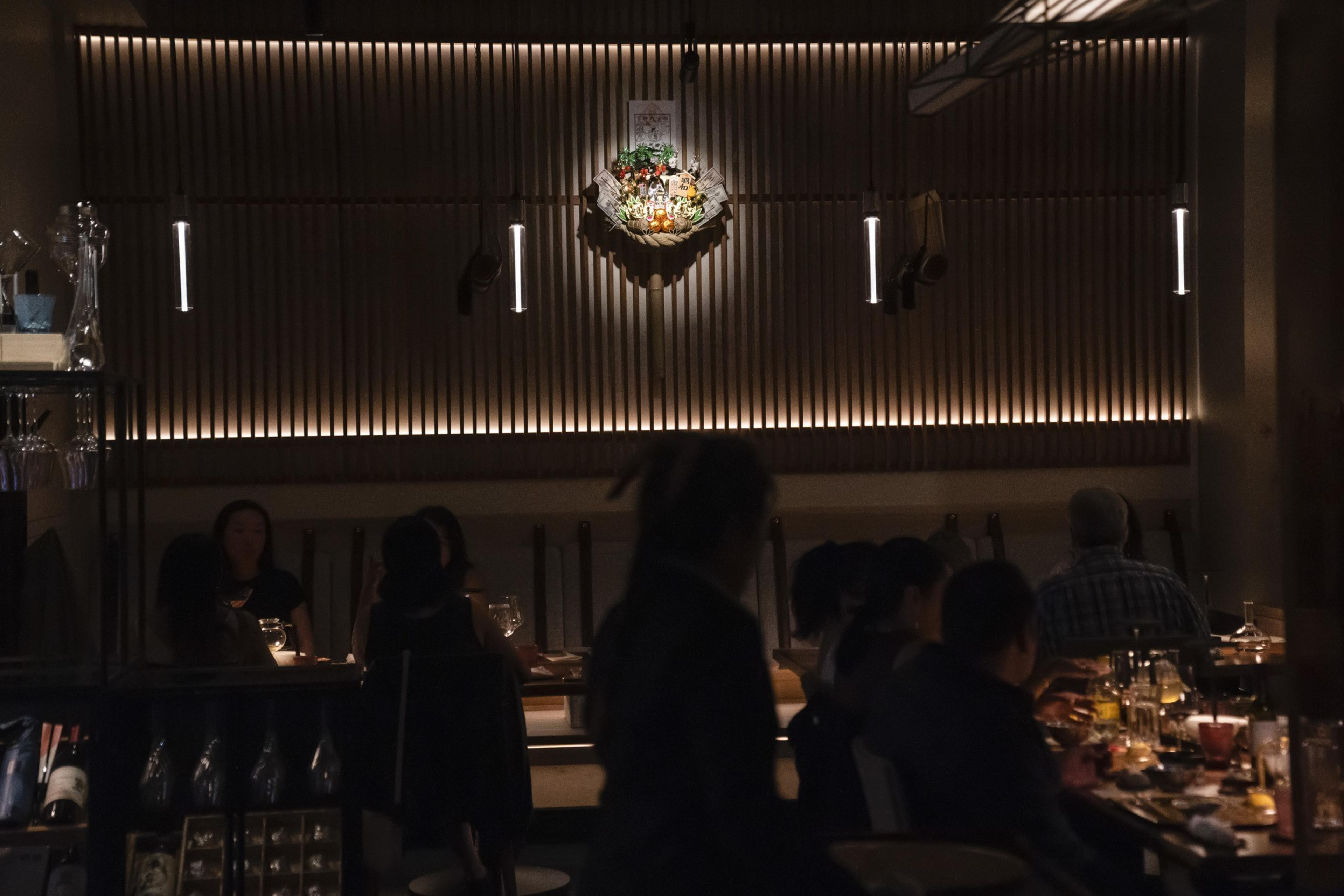 The image depicts a dimly lit restaurant with patrons seated at tables. Warm lighting highlights a decorative wall piece, creating a cozy, intimate atmosphere.