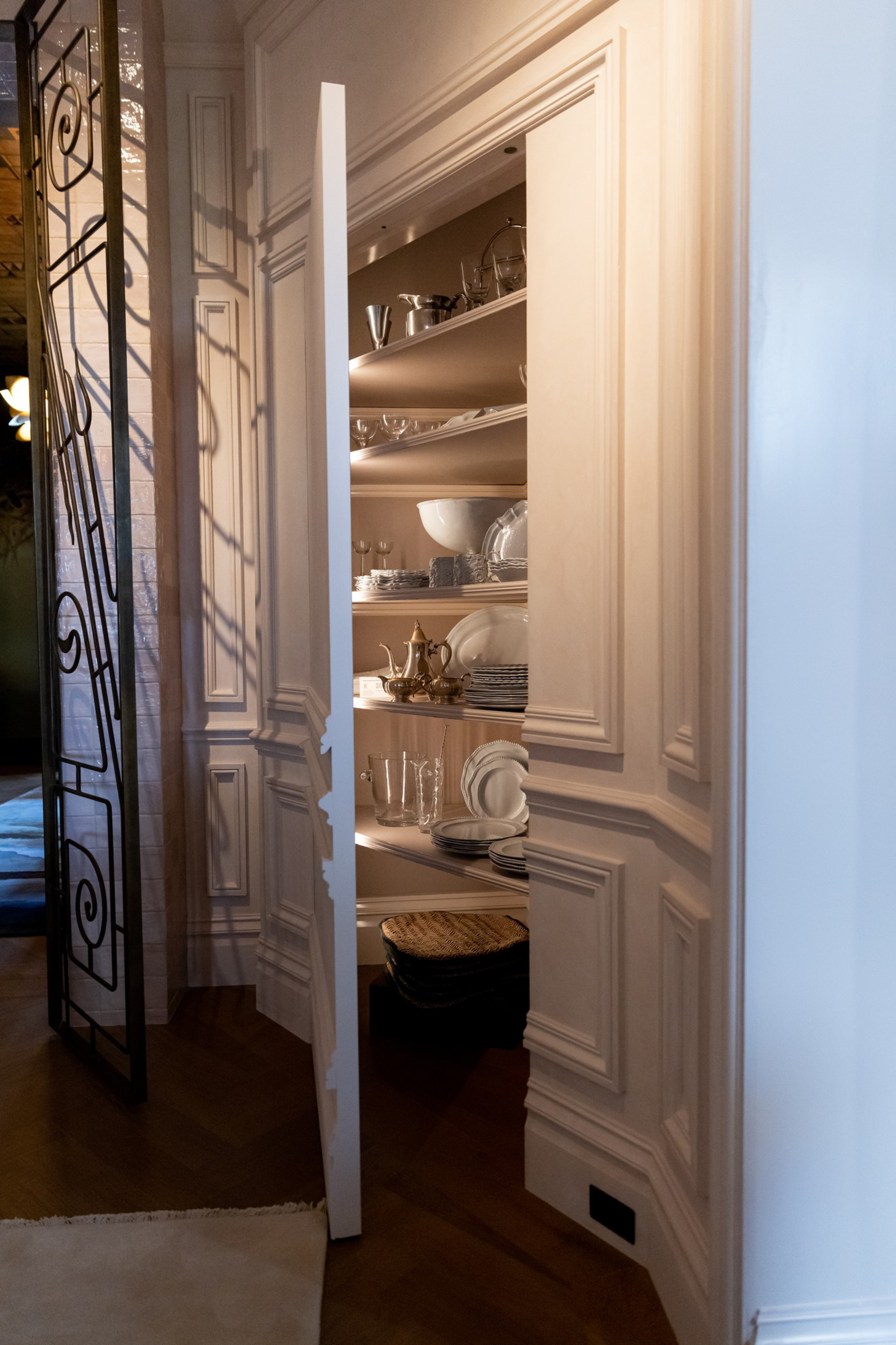 A partially open white paneled door reveals shelves filled with glassware, plates, bowls, and a silver tea set inside a well-lit pantry.