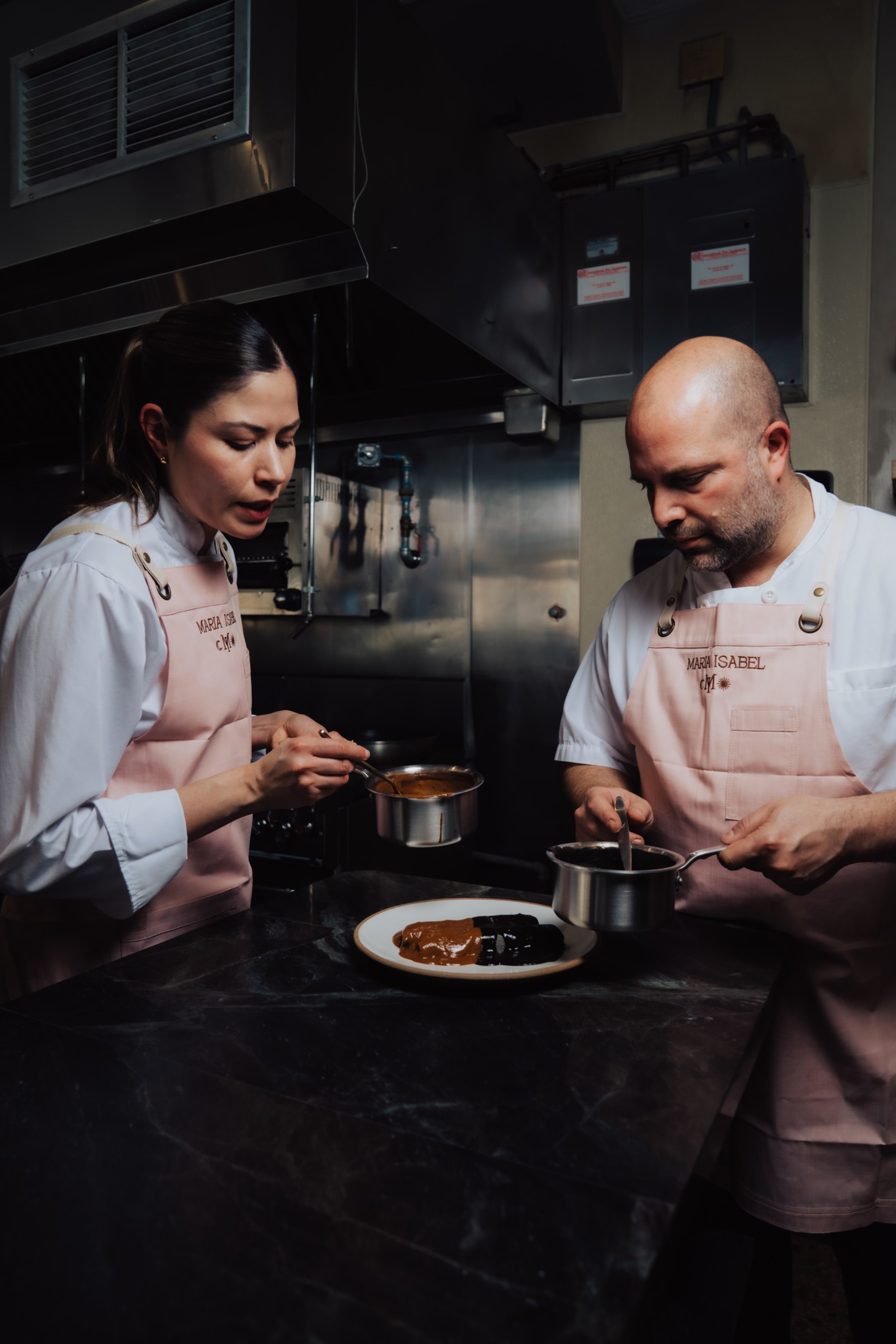 Two chefs in pink aprons carefully pour sauce onto a plate with food in a dimly lit professional kitchen.
