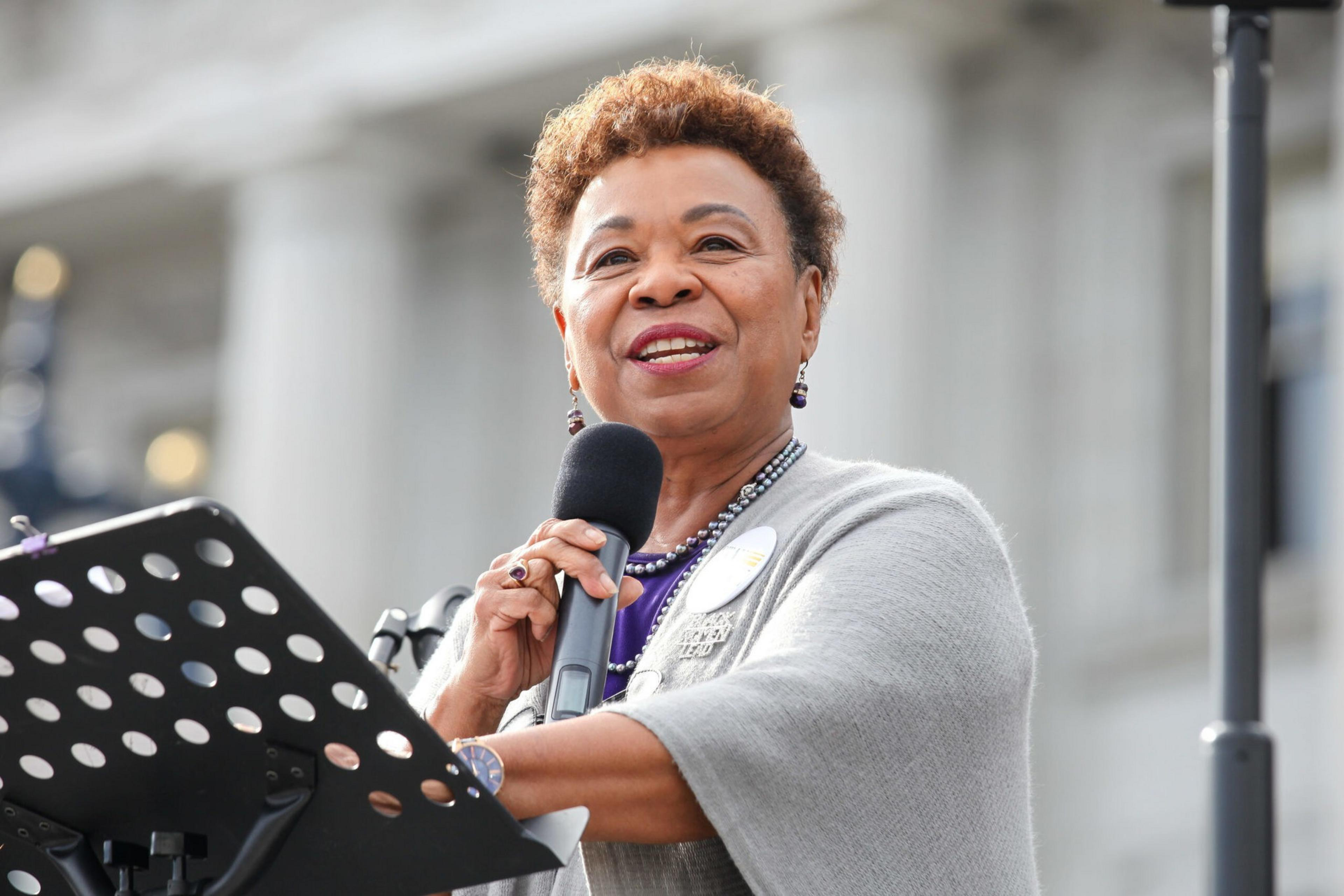 A person is speaking into a microphone at a podium. They are wearing a gray shawl and a round pin, standing in front of a blurred building background.