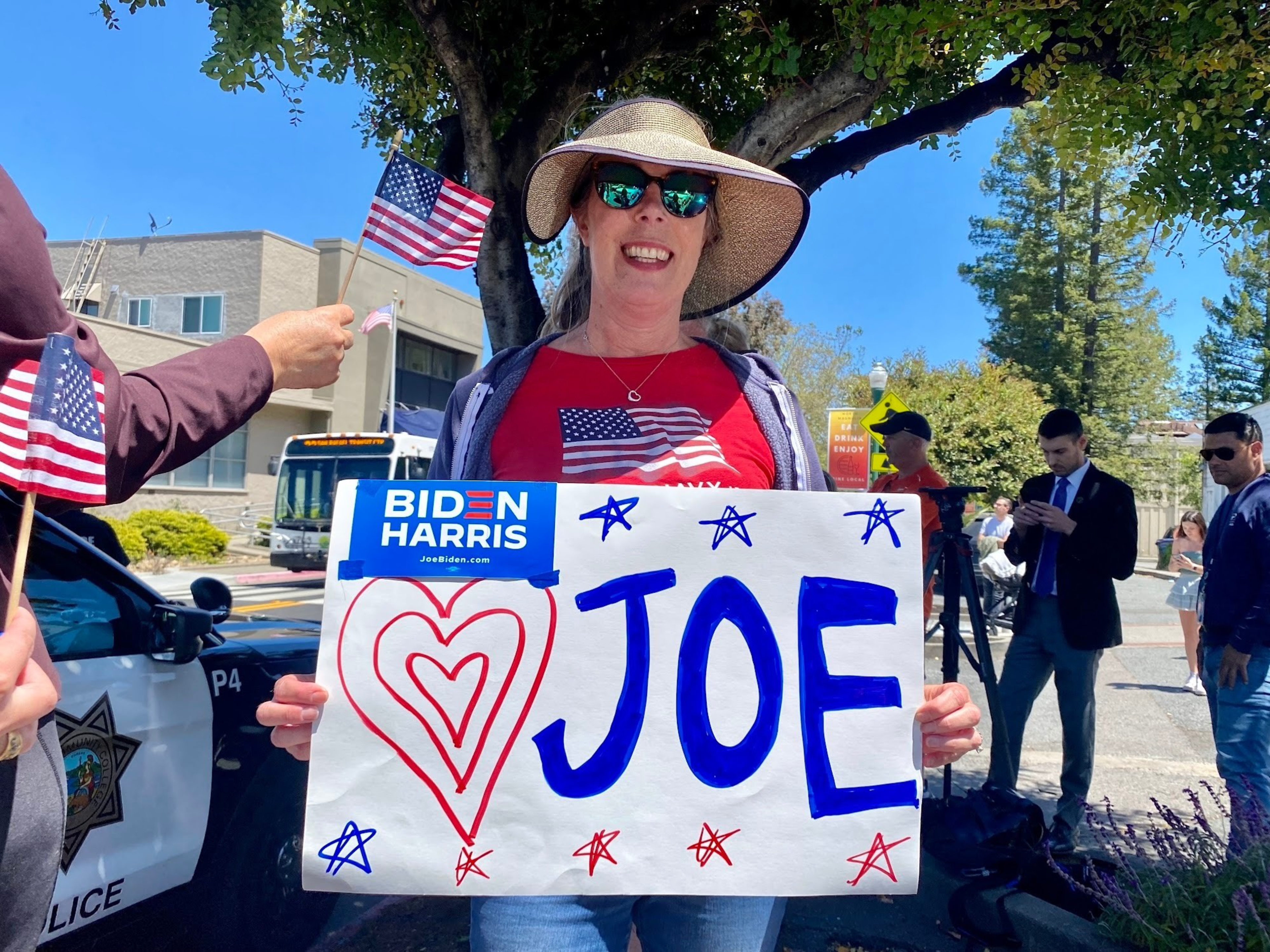 A woman wearing a sunhat and sunglasses holds a "Joe" sign decorated with hearts and stars, along with a Biden Harris sticker, while people and police are nearby.