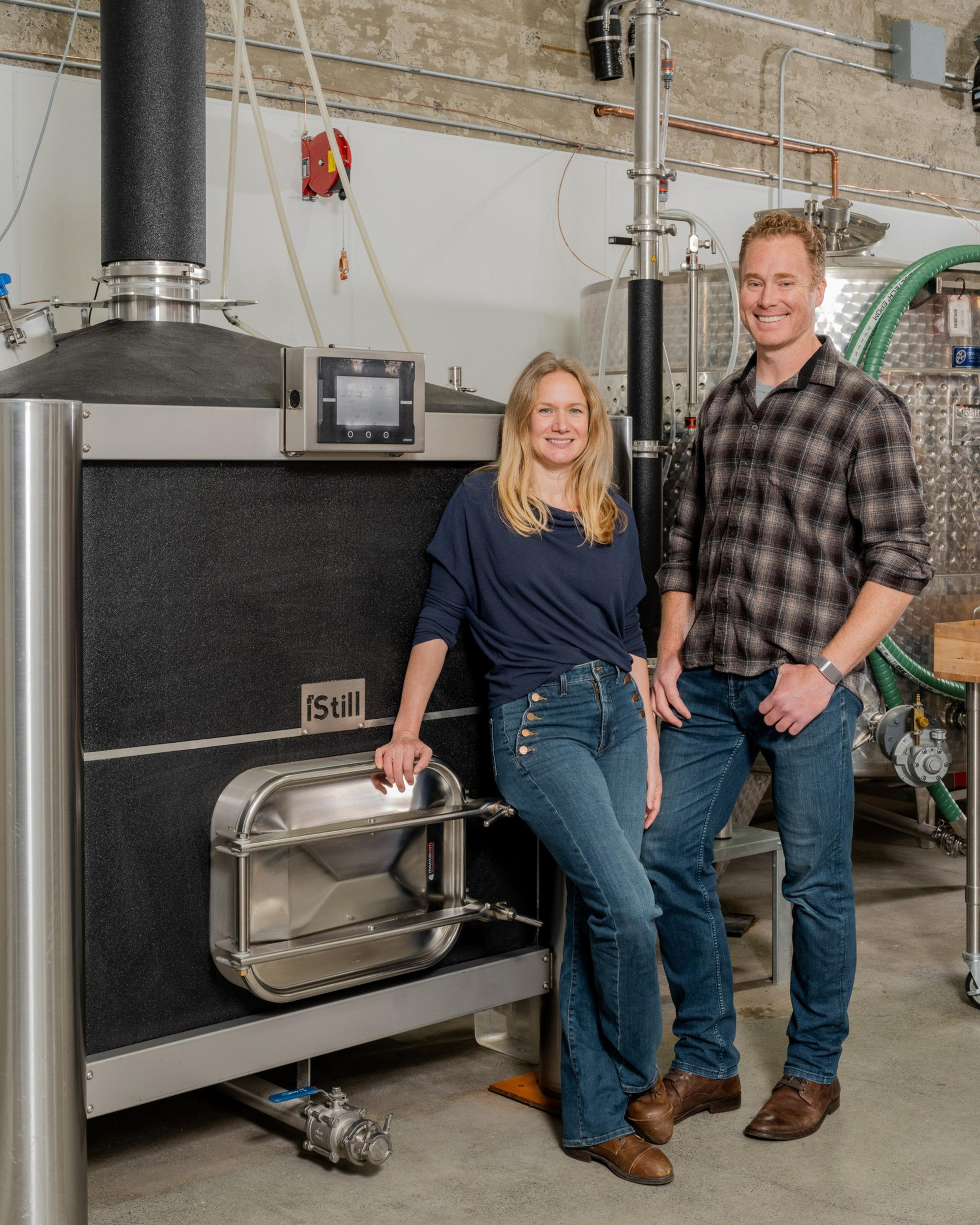 A smiling man and woman stand in a distillery, leaning on large black and stainless steel equipment labeled “Still.”