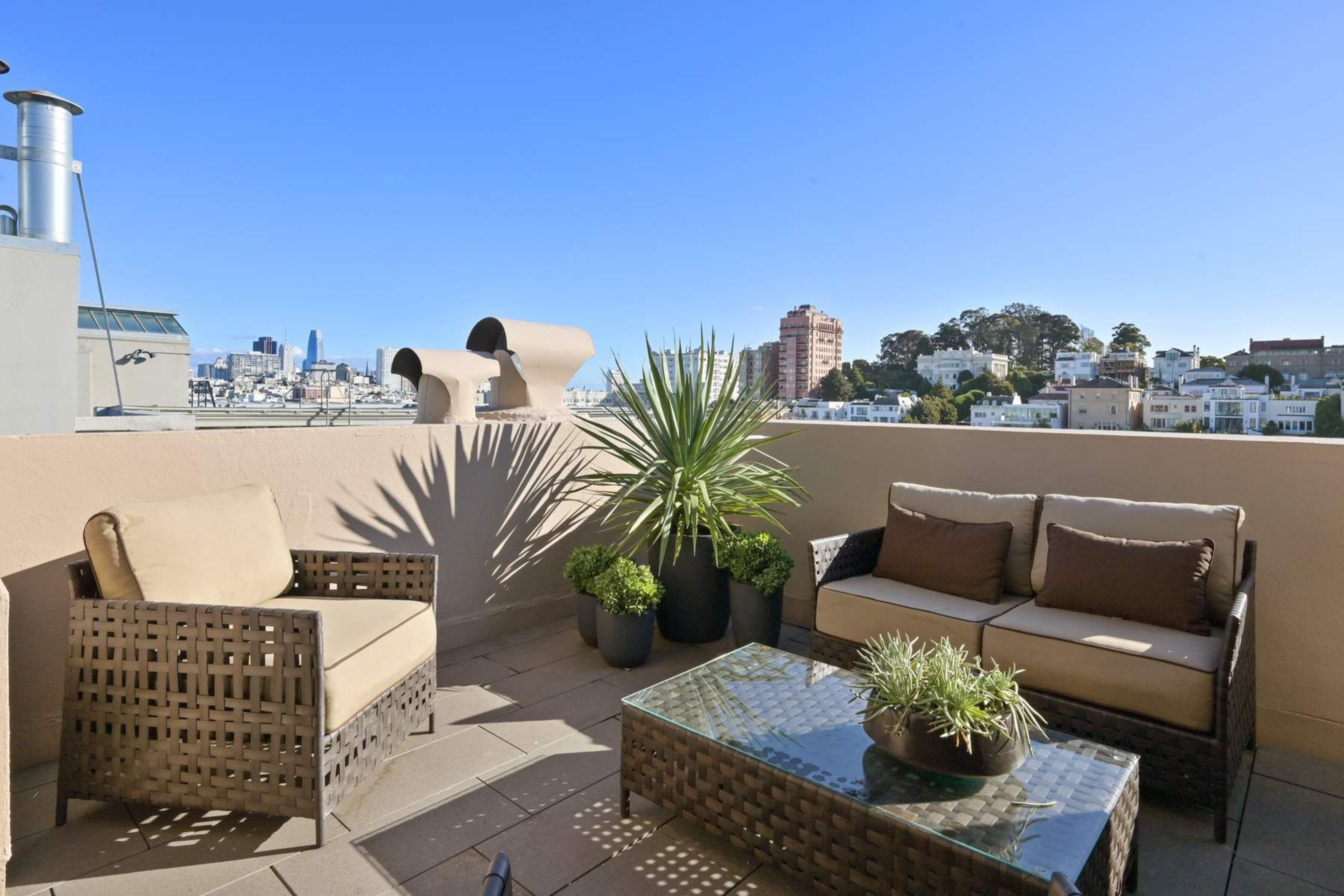 A rooftop patio with woven furniture, including a sofa, chair, glass-top coffee table, and potted plants, overlooking a cityscape under clear blue sky.