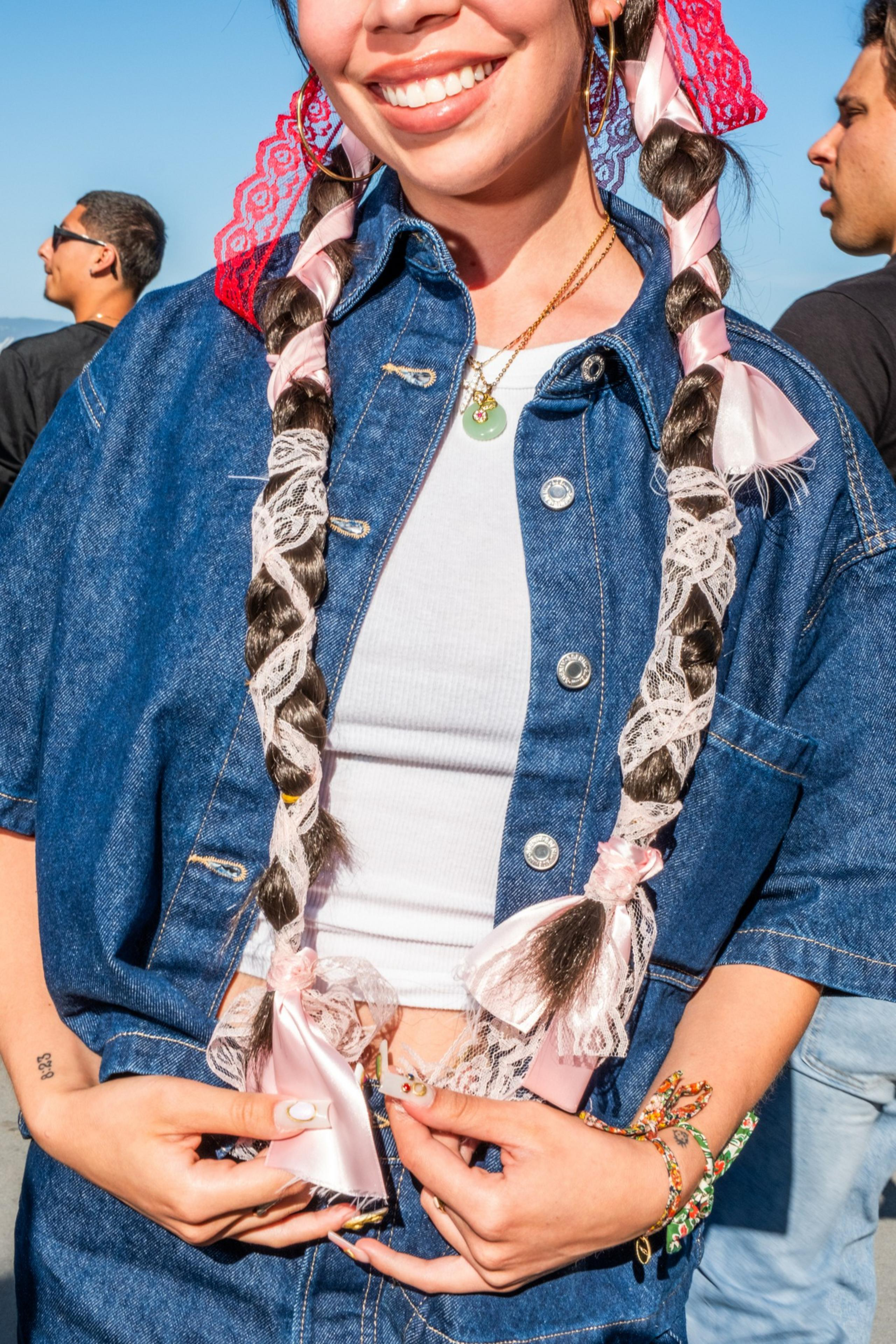 A smiling person with long braids wrapped in pink ribbon and lace, wearing a denim jacket over a white top, accessorized with necklaces and bracelets.