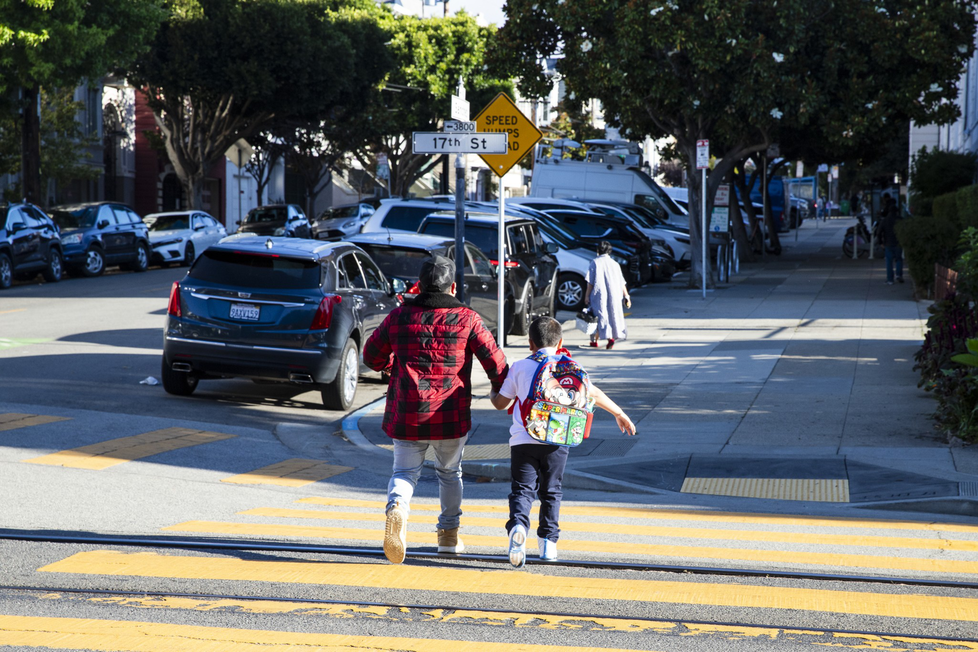 A man in a red jacket and a child with a colorful backpack cross a sunlit street at a crosswalk near a sign reading "17th St" and "Speed Humps Ahead."
