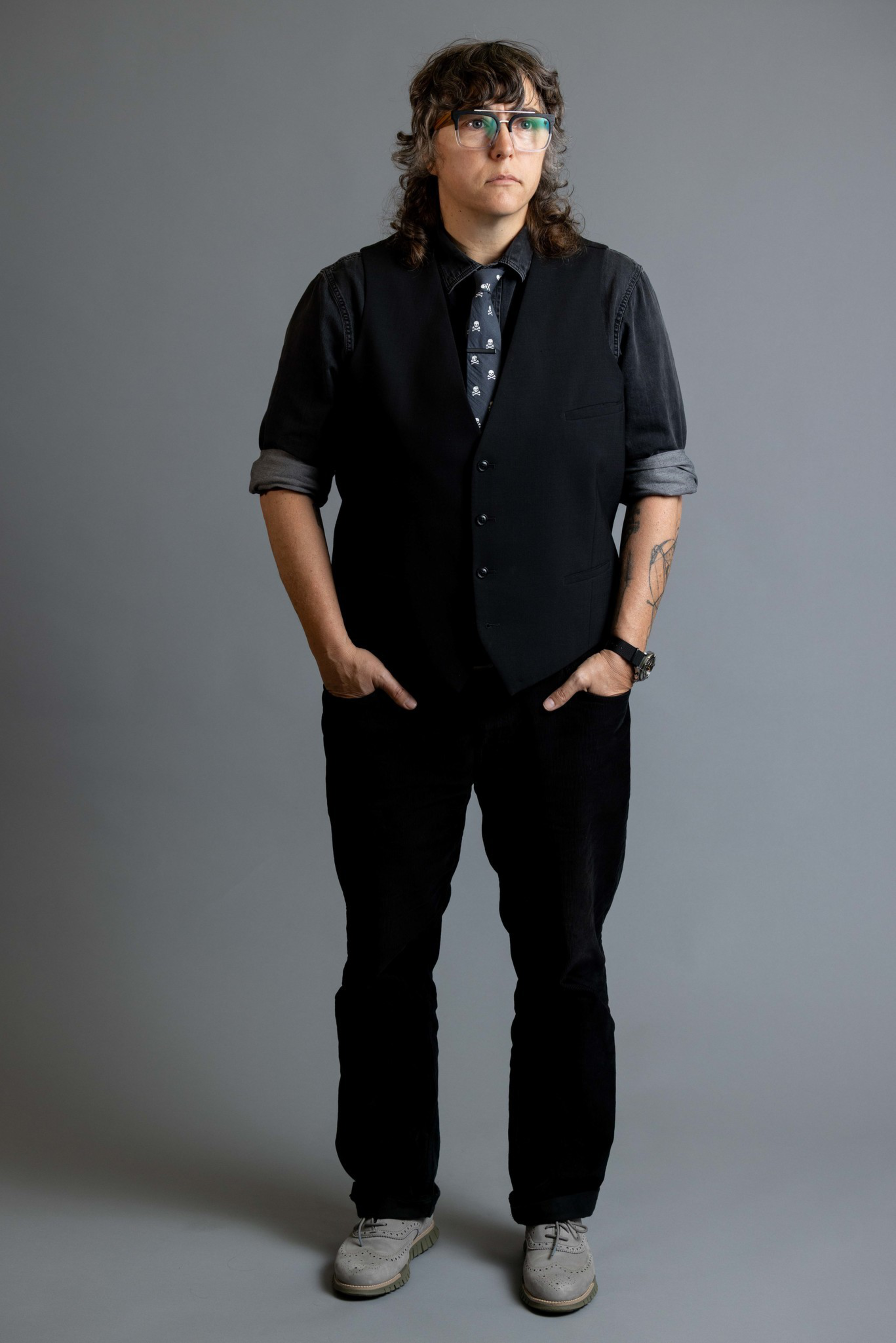A person with curly hair and glasses wears a black vest over a rolled-up sleeve shirt, a patterned tie, black pants, and white shoes, standing with hands in pockets.
