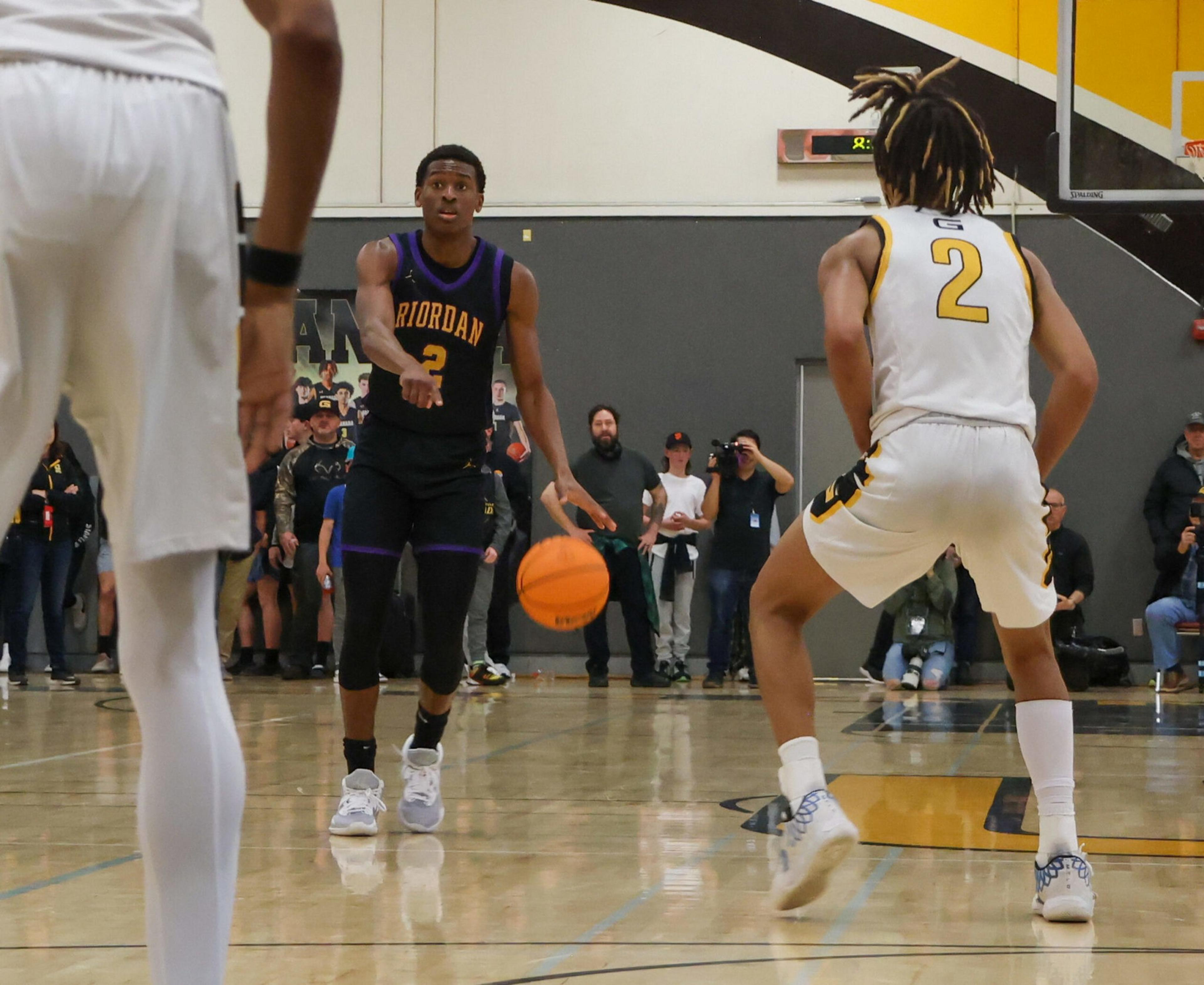 A basketball player in a black uniform passes the ball while being defended by two players in white uniforms on an indoor court.