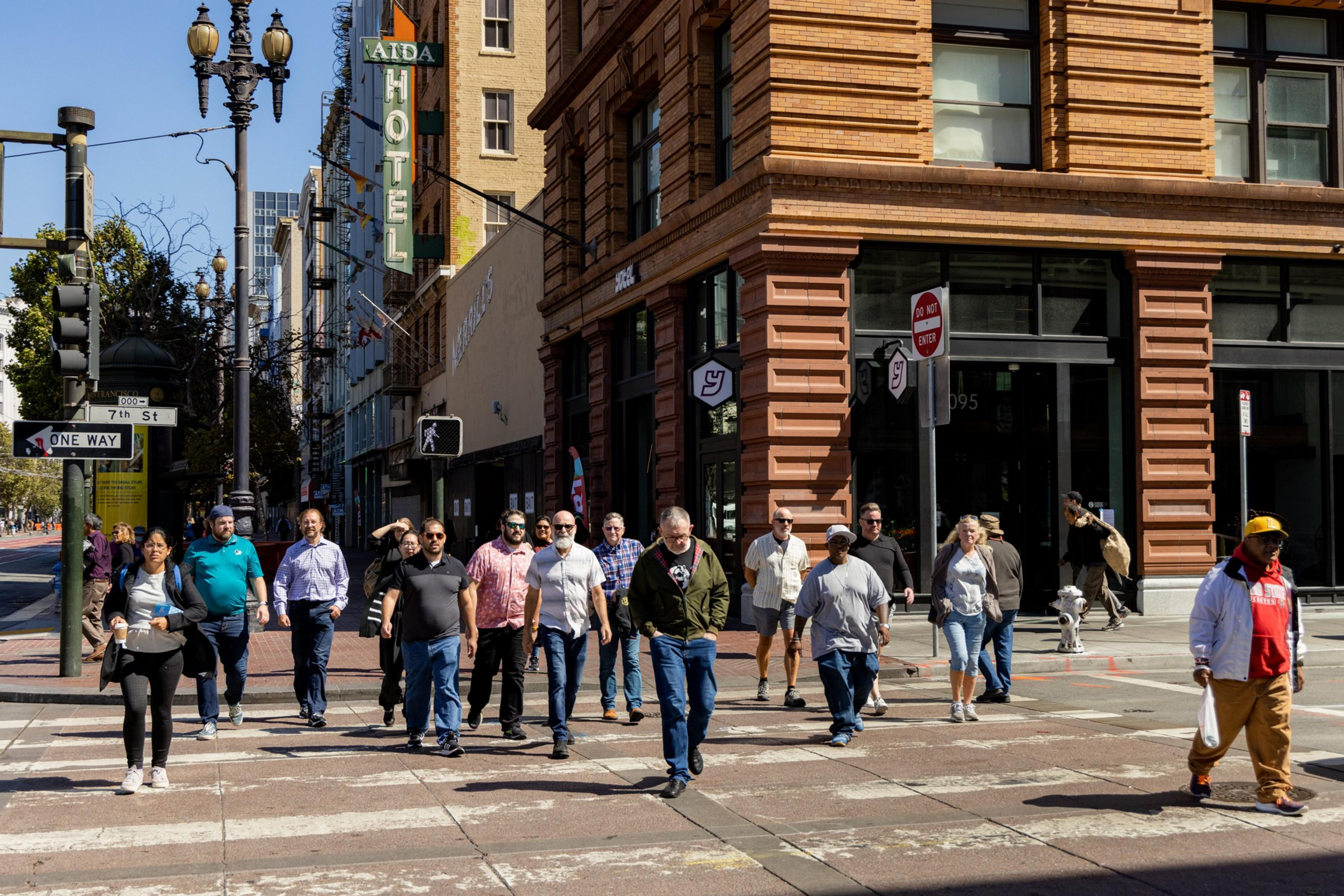 A group of people is crossing a city street. They are walking away from a corner building with a large sign that reads "Aida Hotel." The scene is sunny and urban.