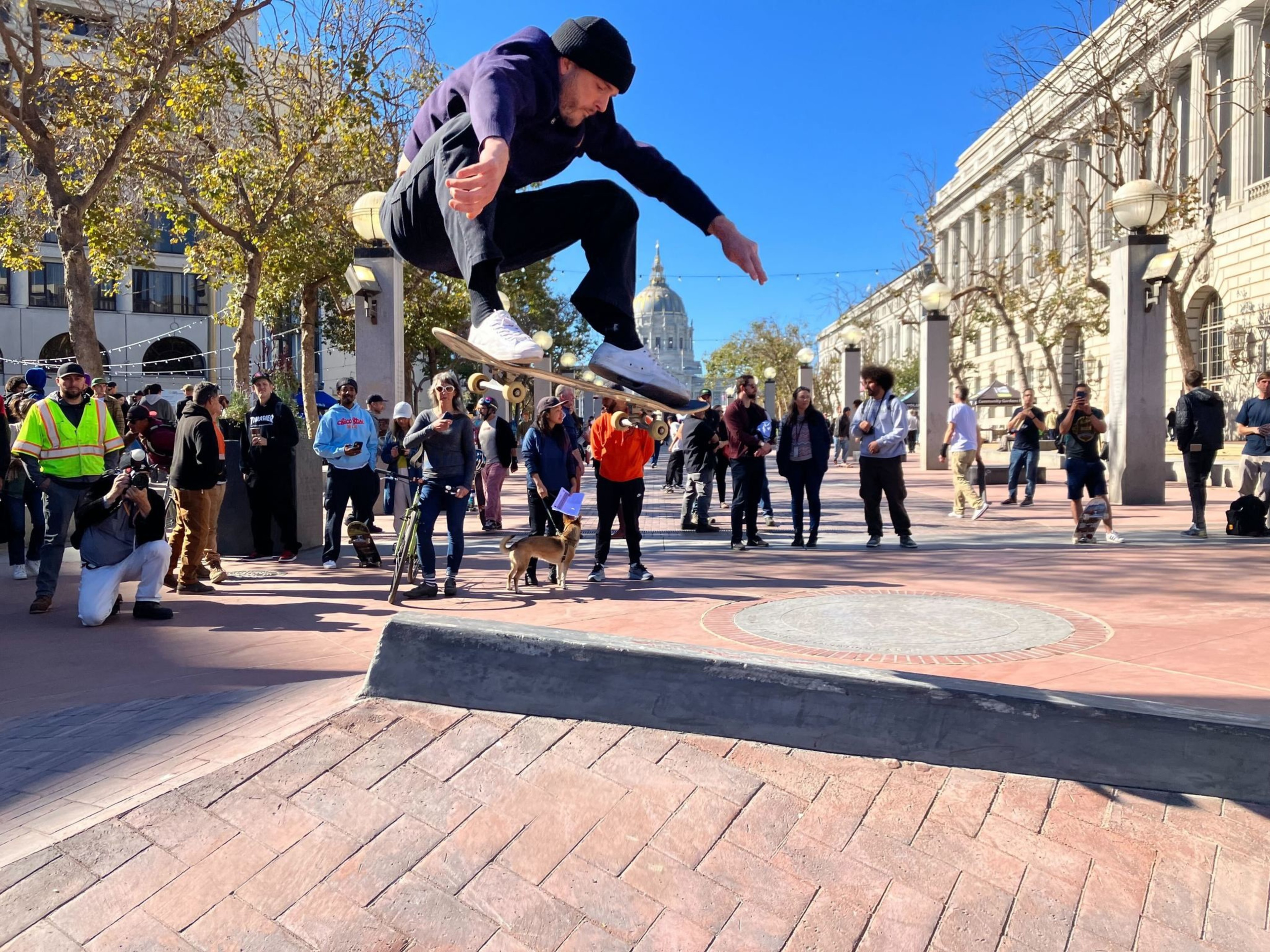 A man on a skateboard, wearing a black beanie, a purple hoodie, black jeans and white sneakers, hoovers in the air in a crouched position over a brick and concrete skate park feature while onlookers watch in the background.