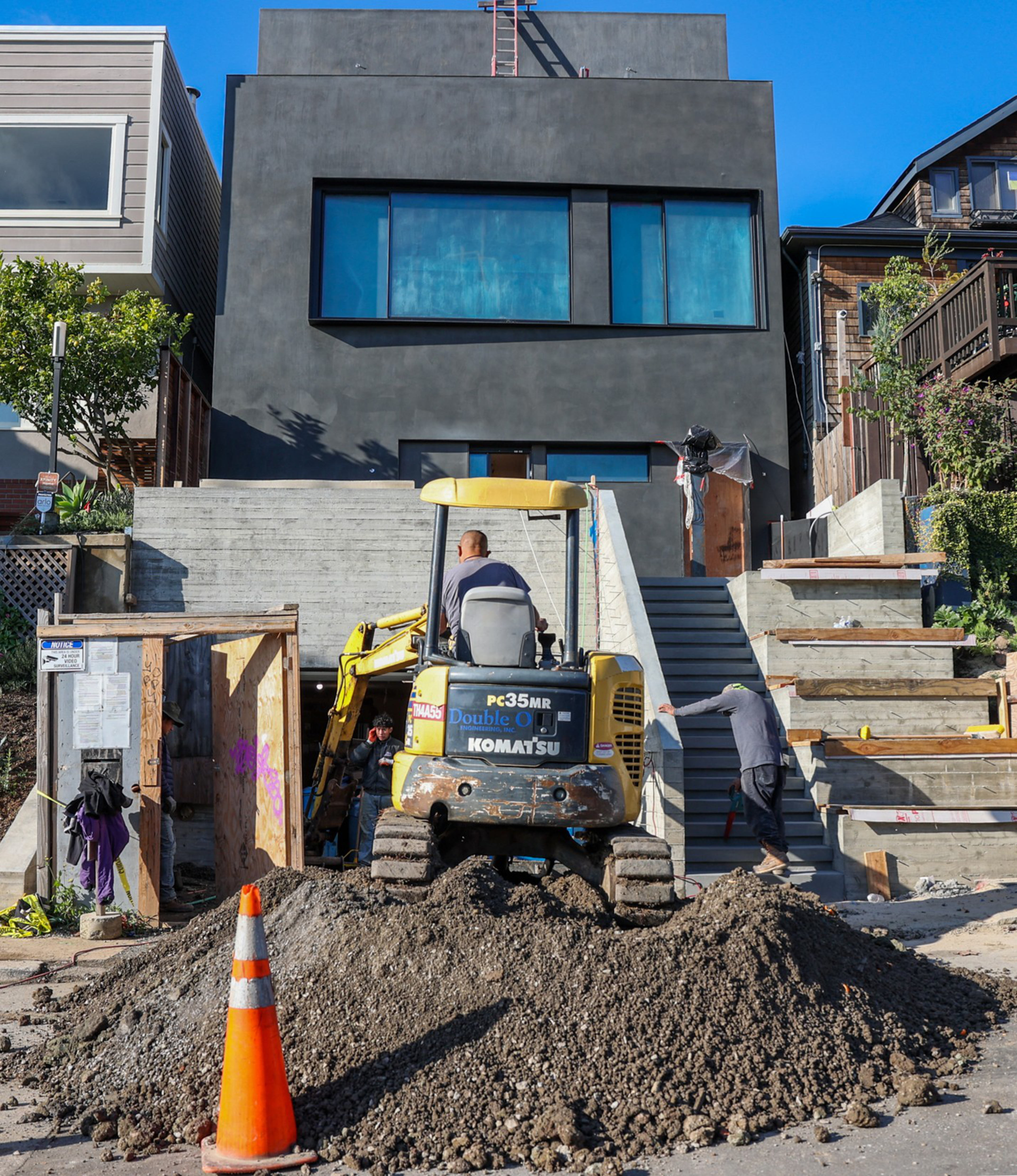 A mini excavator on a pile of dirt, with a worker behind it, at a modern home construction site.
