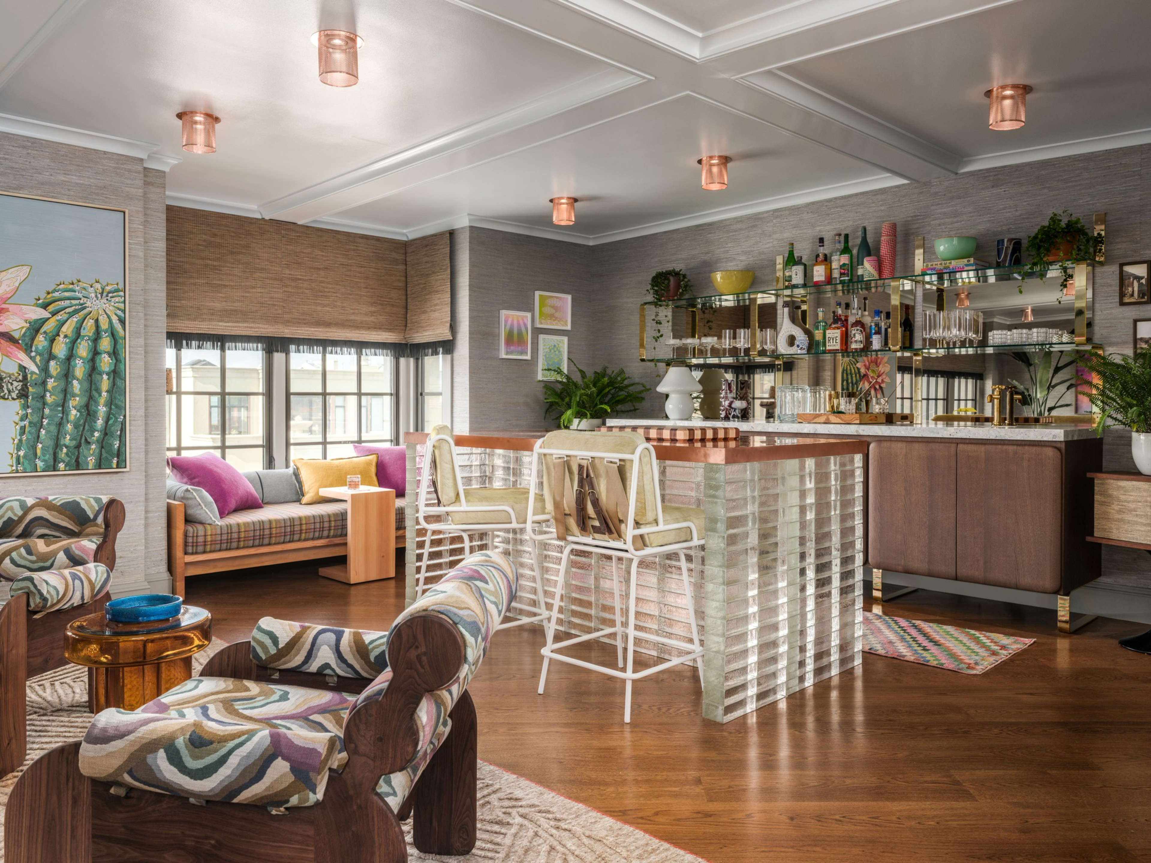 A cozy living area with patterned armchairs, a wooden bar with glass block sides, bar stools, a window seat with pillows, and plants near a mirrored bar wall.