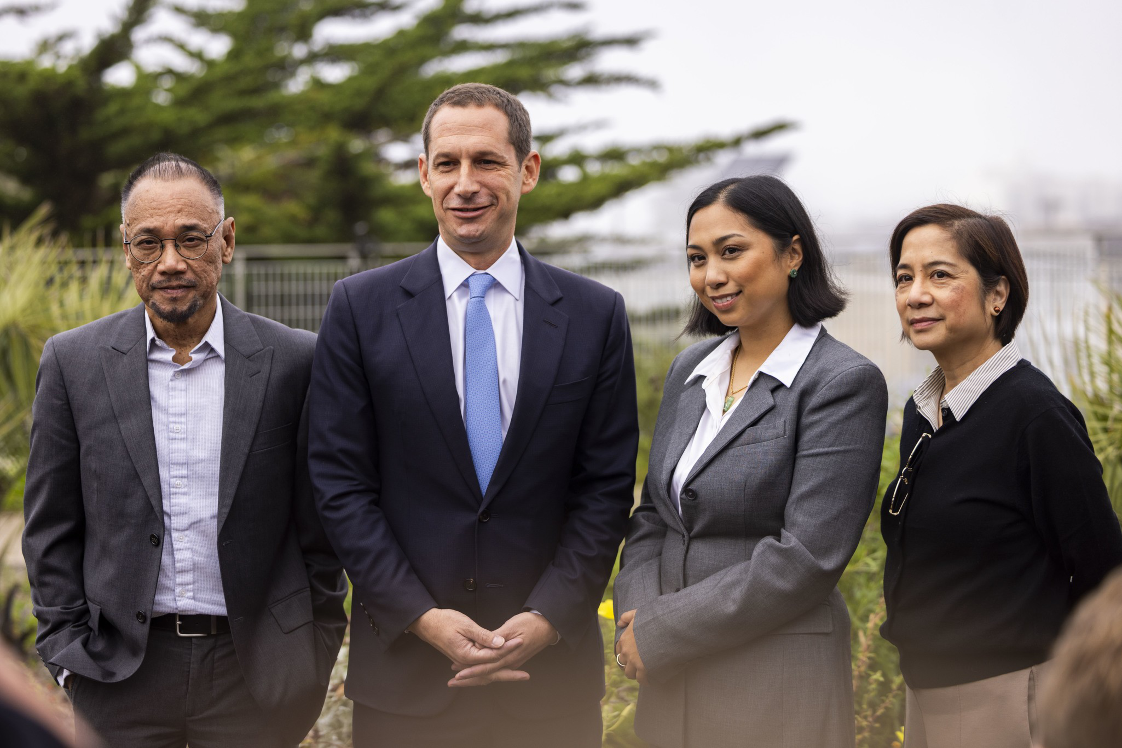 Four professionally dressed people stand outdoors with plants and trees in the background, smiling and posing for a photo.