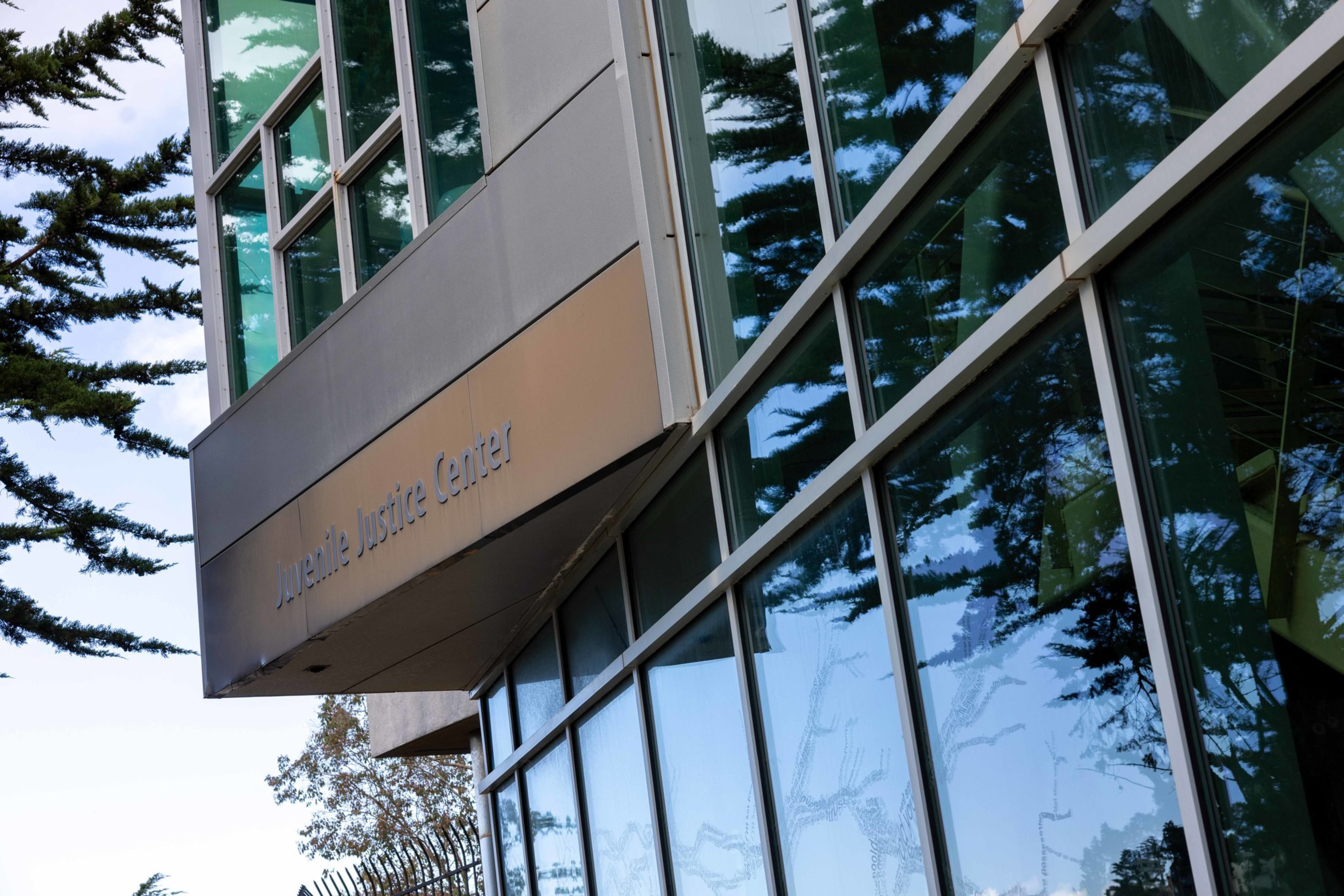 A modern building with large glass windows reflecting trees and a sign reading “Juvenile Justice Center.”