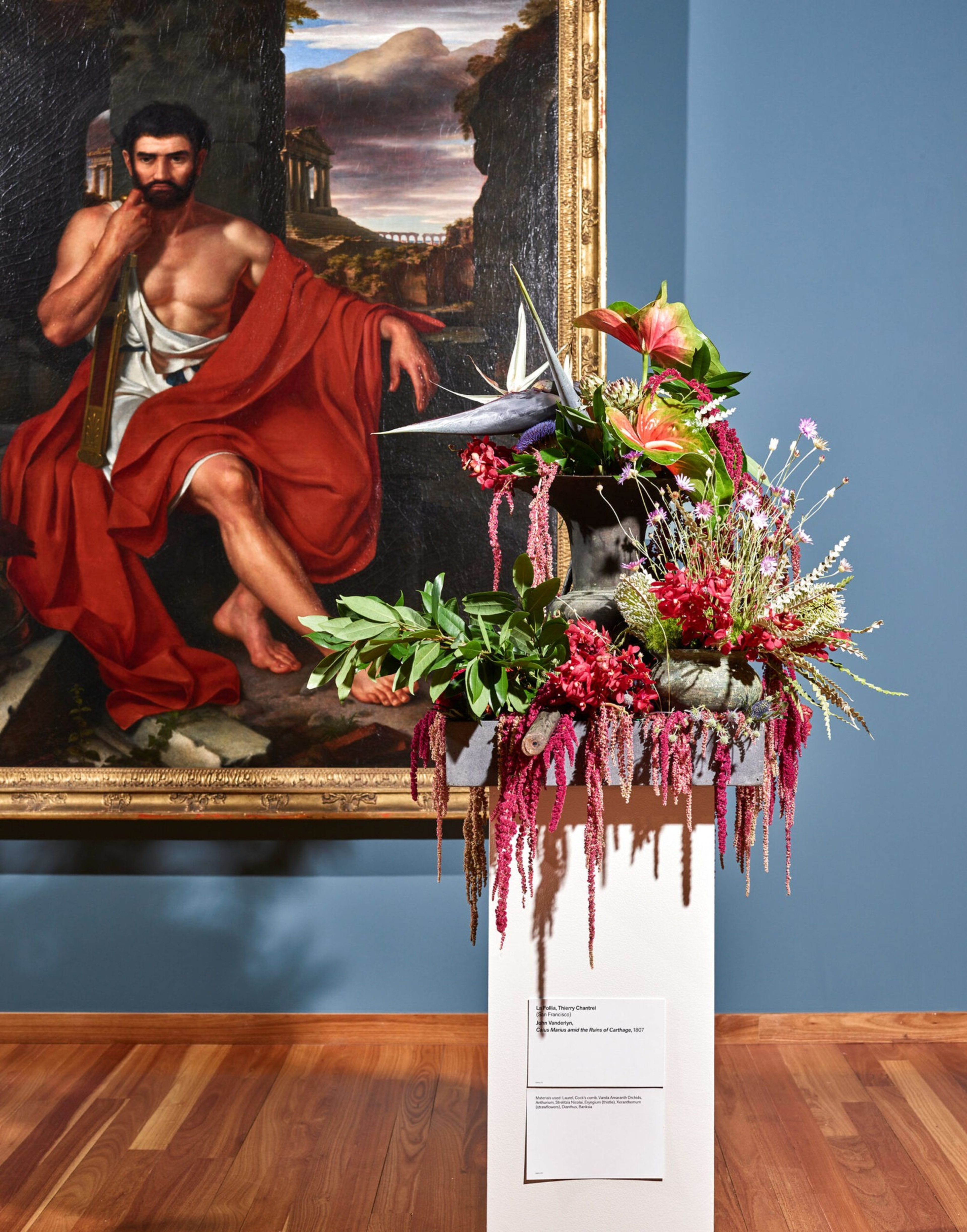 A classical painting of a seated man draped in red cloth hangs behind a pedestal holding a vibrant floral arrangement with pink, green, and purple flowers.