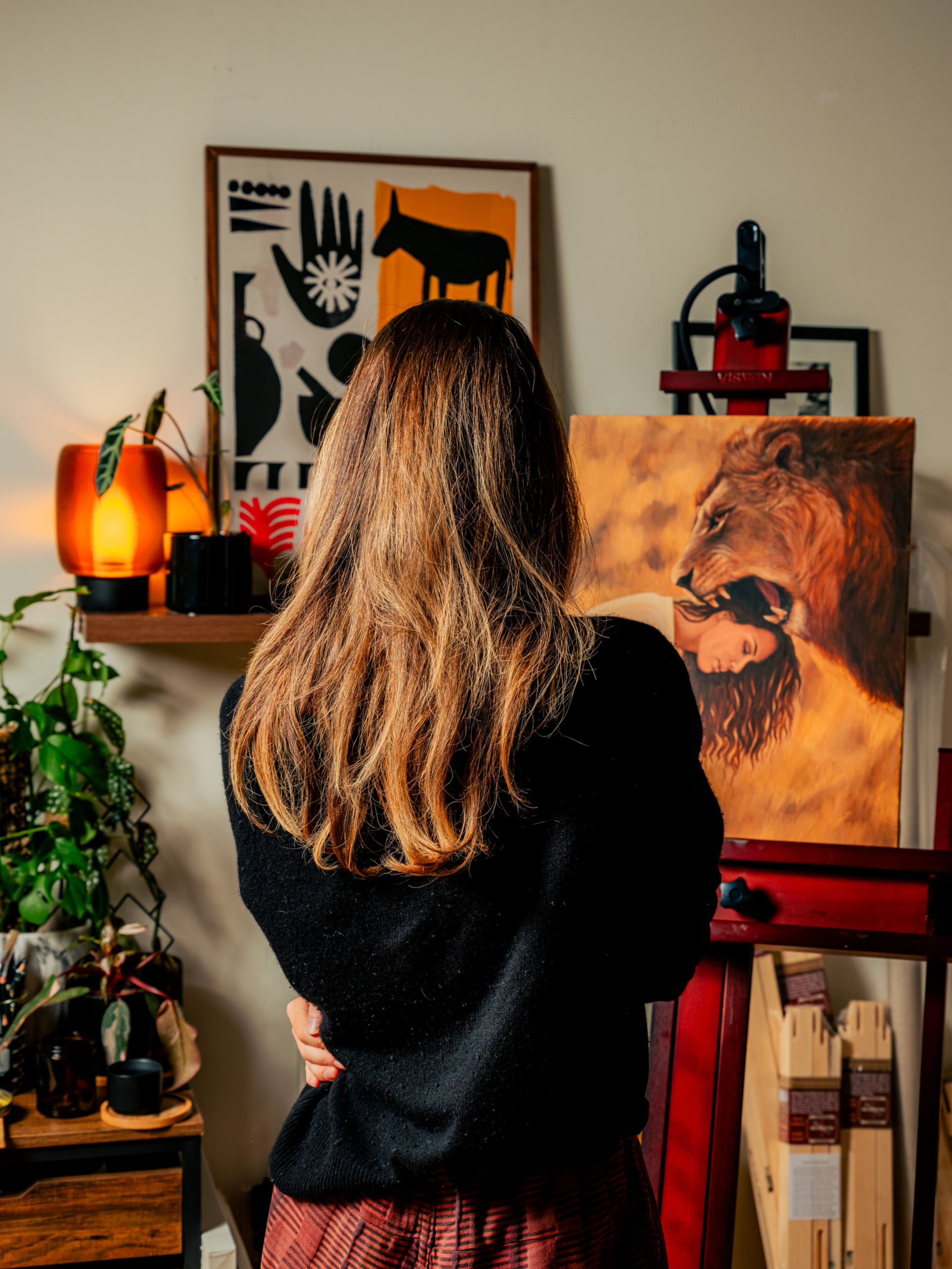 A woman with long brown hair stands facing an easel with a painting of a lion and a woman’s face merging, surrounded by plants and abstract framed art.