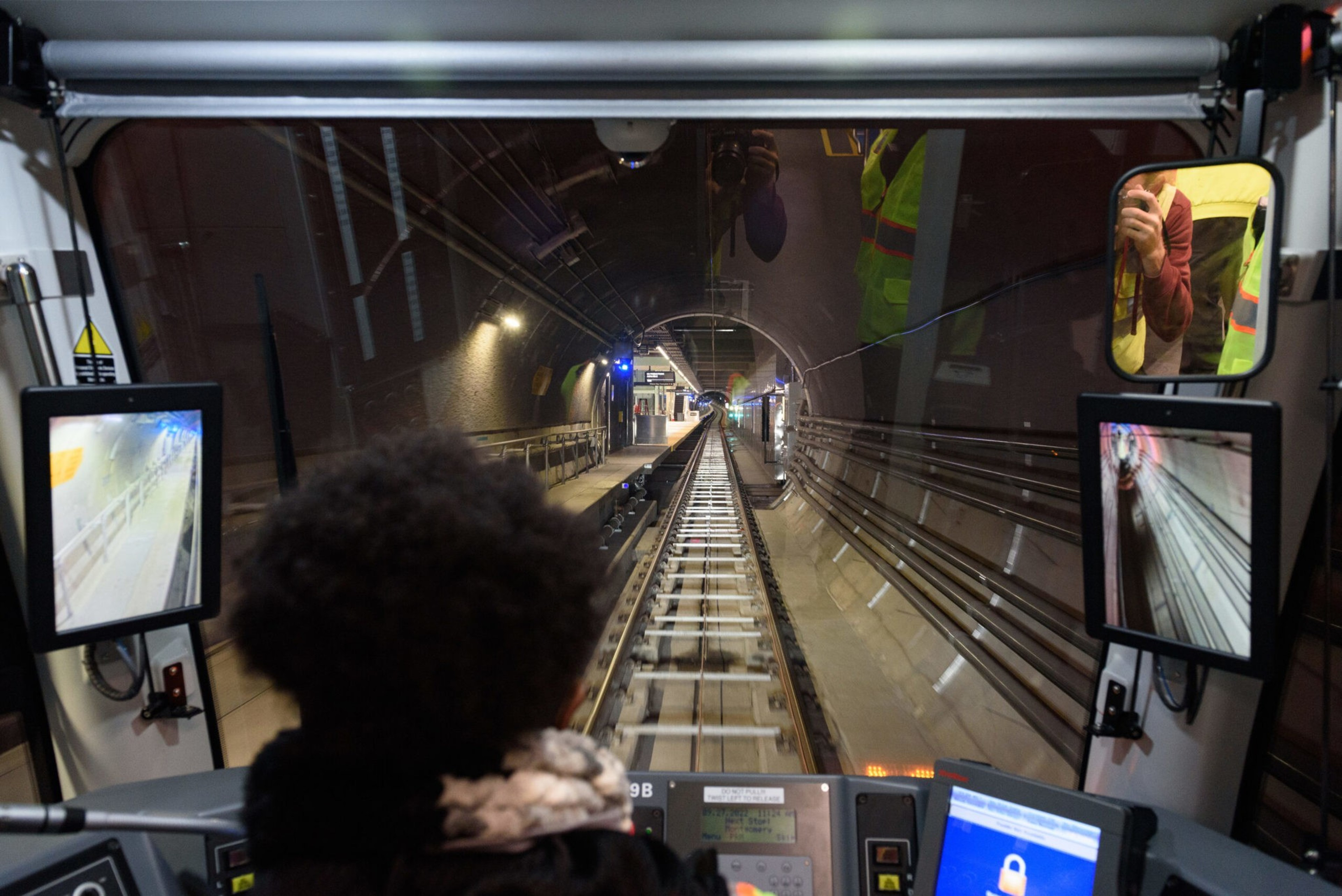 A person operates a train in a tunnel, viewing tracks ahead through large windows and two screens showing live camera feeds from the train's sides.