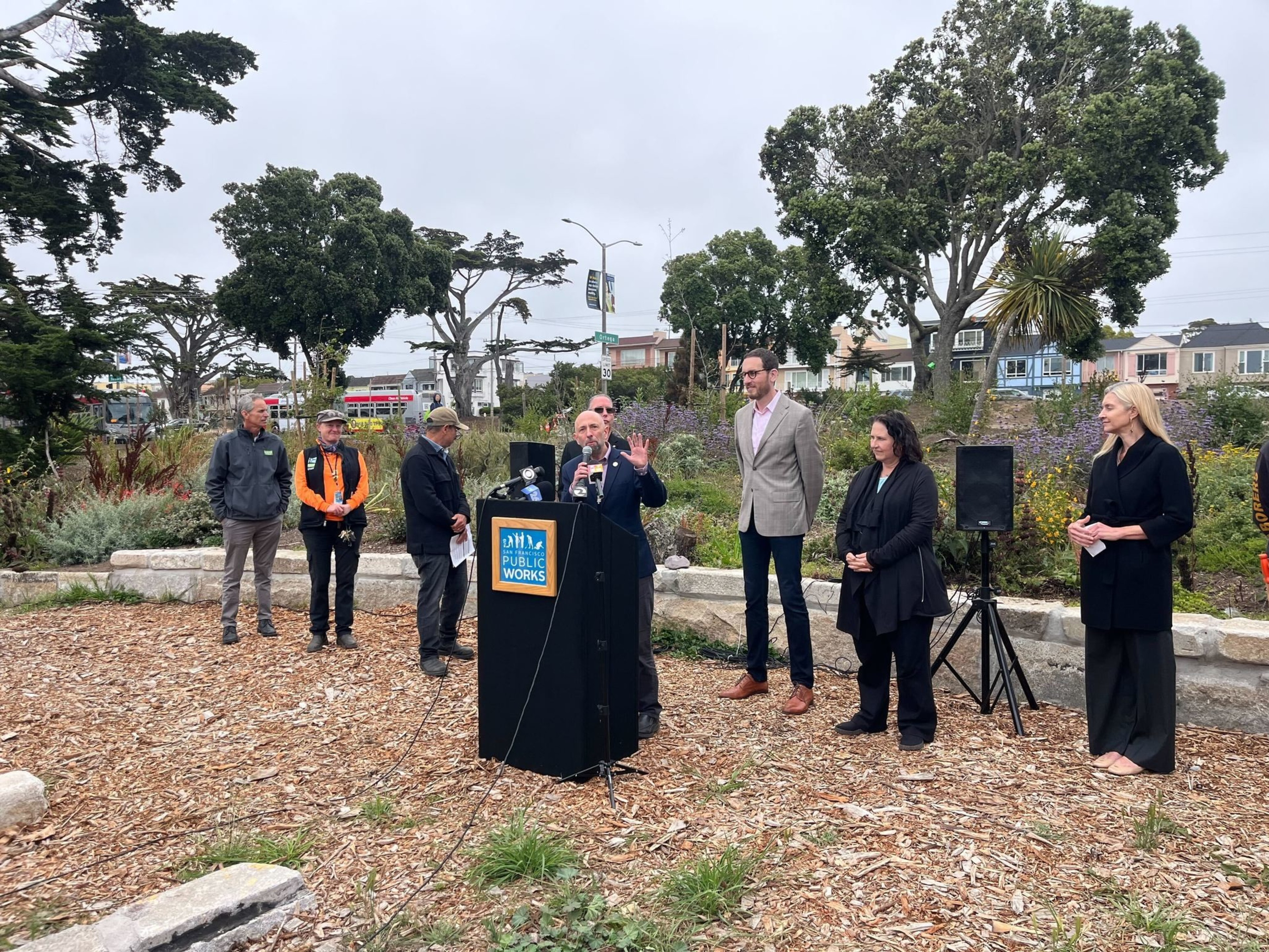 A group of people stand outdoors near a podium labeled &quot;Public Works.&quot; Trees and greenery are in the background, with some wearing professional attire and others in casual wear.
