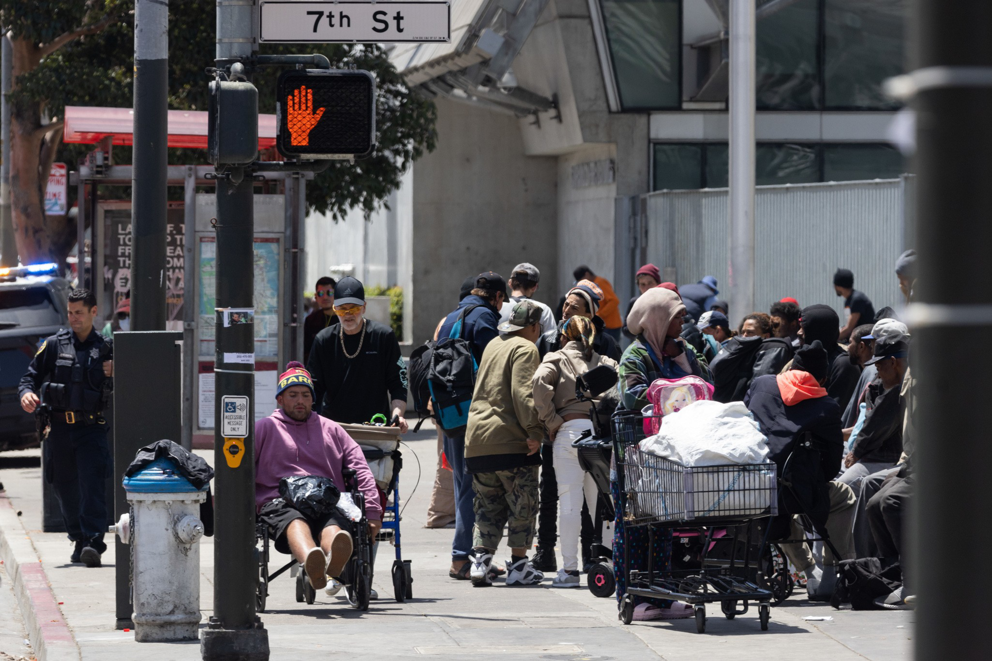 A group of people congregate next to bus stop next to a chain link fence with a street sign that reads 7th St.