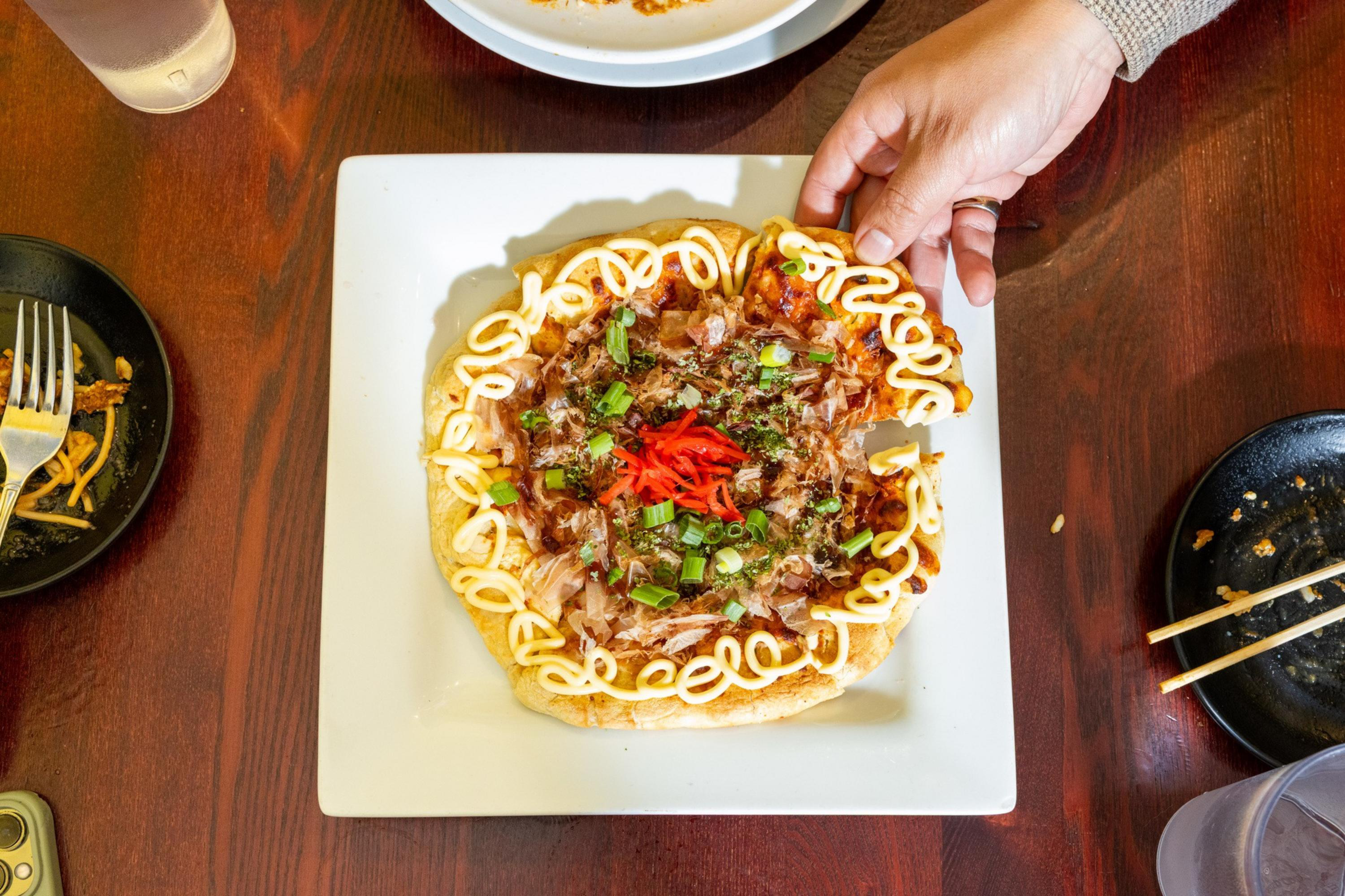 A hand is lifting a slice from a round Japanese-style pizza topped with mayonnaise, green onions, red pickled ginger, and bonito flakes, on a white square plate.
