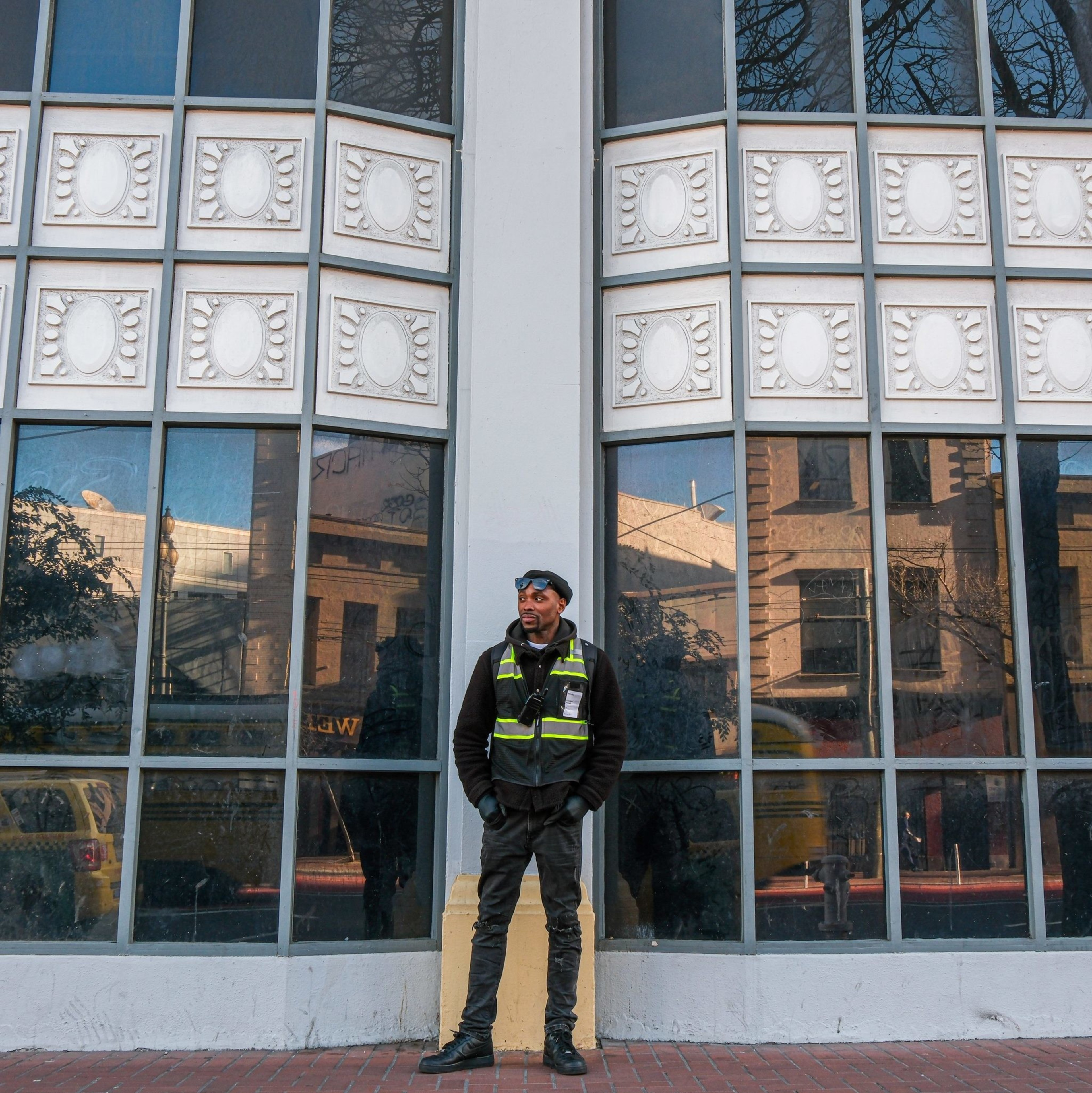 A person stands in front of tall, reflective windows, wearing a dark uniform and a high-visibility vest. The background shows a building facade and tree reflections.