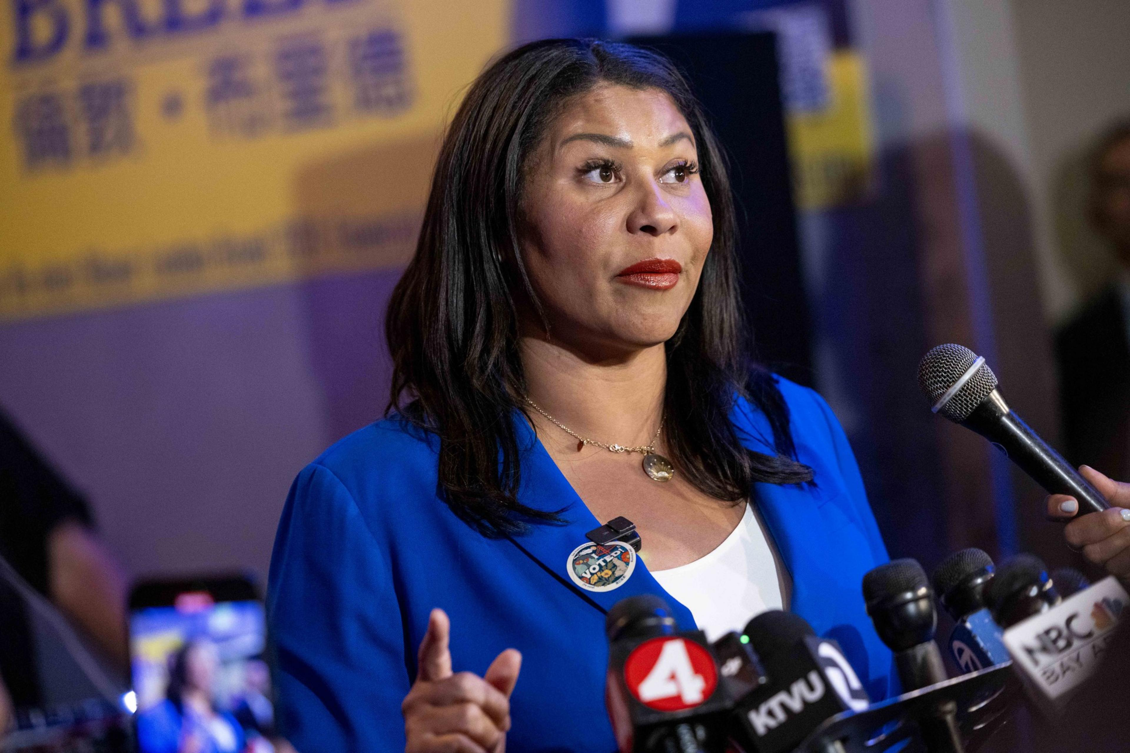 A woman in a blue blazer speaks at a podium with several microphones in front of her. The background features a yellow and purple banner with text.