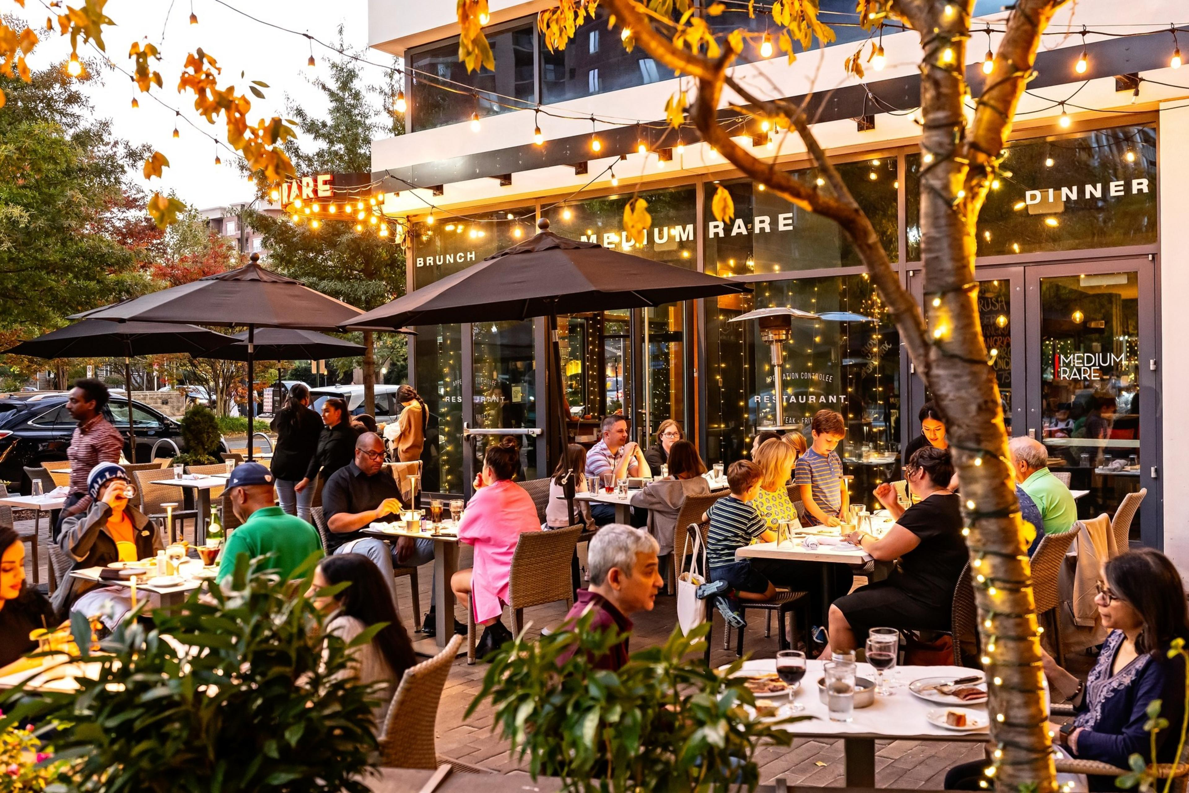About two dozen people sit at tables outside a restaurant near bushes and a tree with Christmas lights wrapped around its trunk.