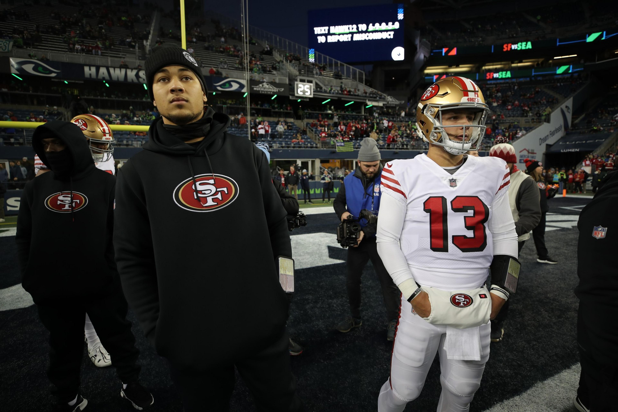 Several San Francisco 49ers players stand on the field before a game, with one player in full white uniform and others in black hoodies, surrounded by photographers.