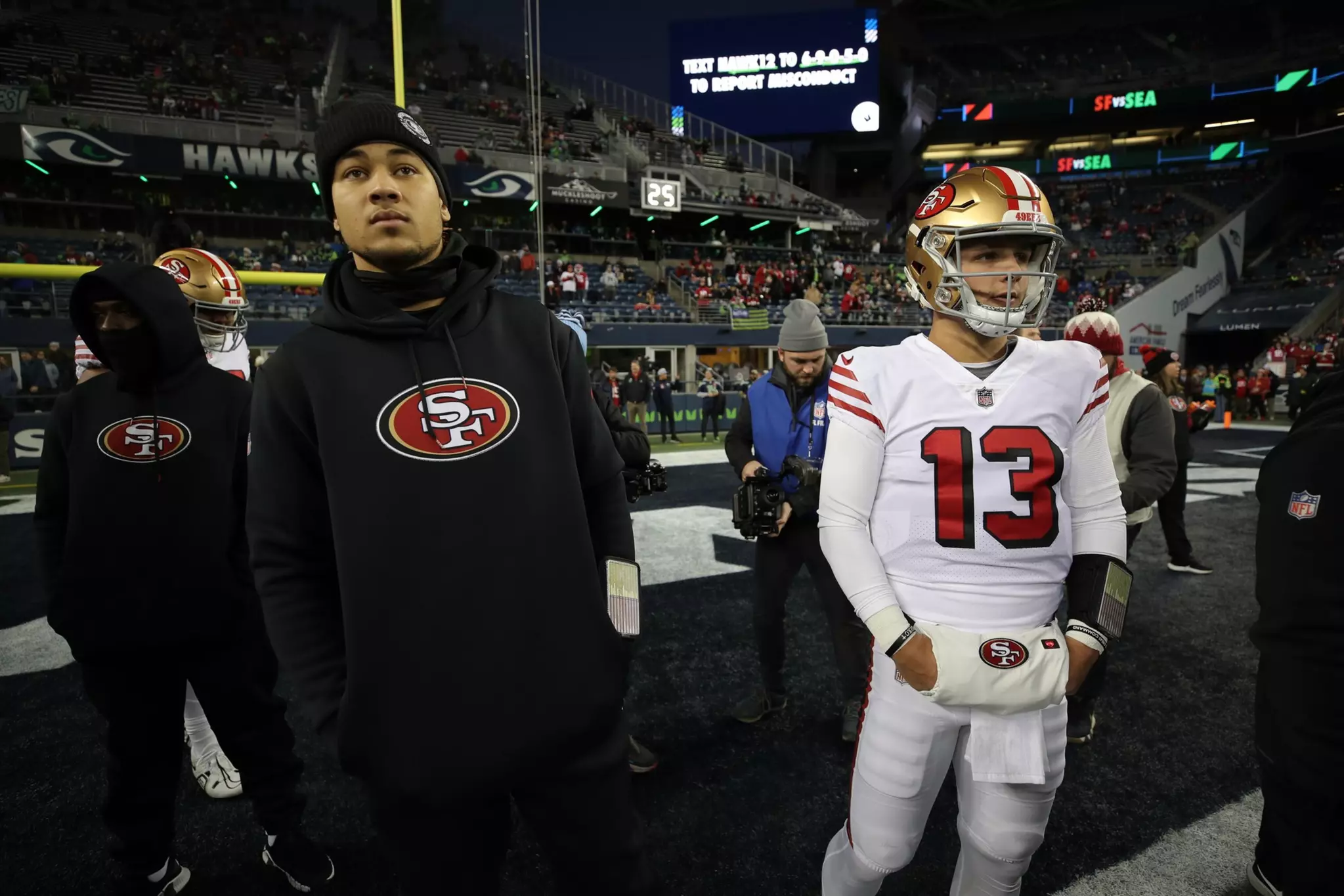 Several San Francisco 49ers players stand on the field before a game, with one player in full white uniform and others in black hoodies, surrounded by photographers.