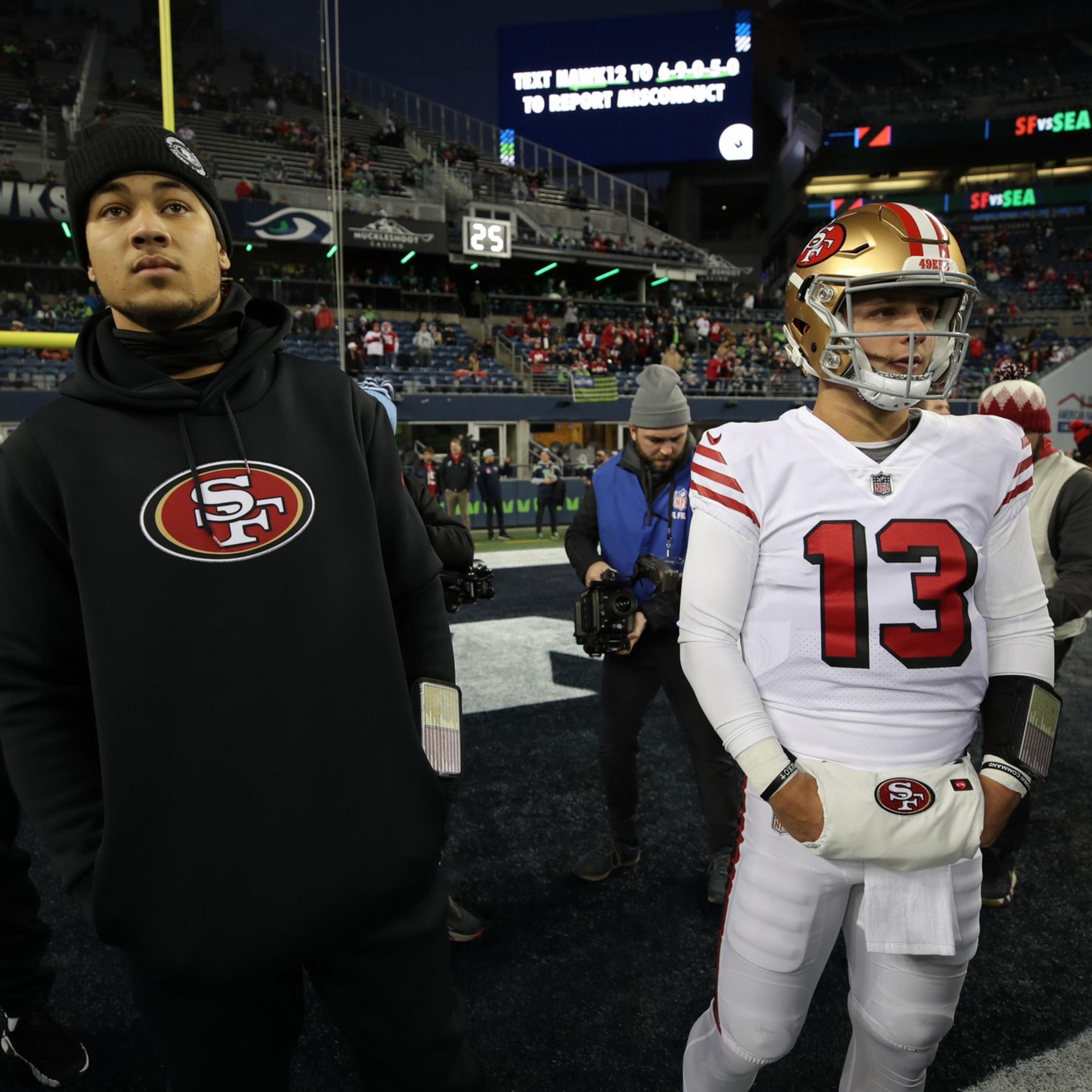 Several San Francisco 49ers players stand on the field before a game, with one player in full white uniform and others in black hoodies, surrounded by photographers.