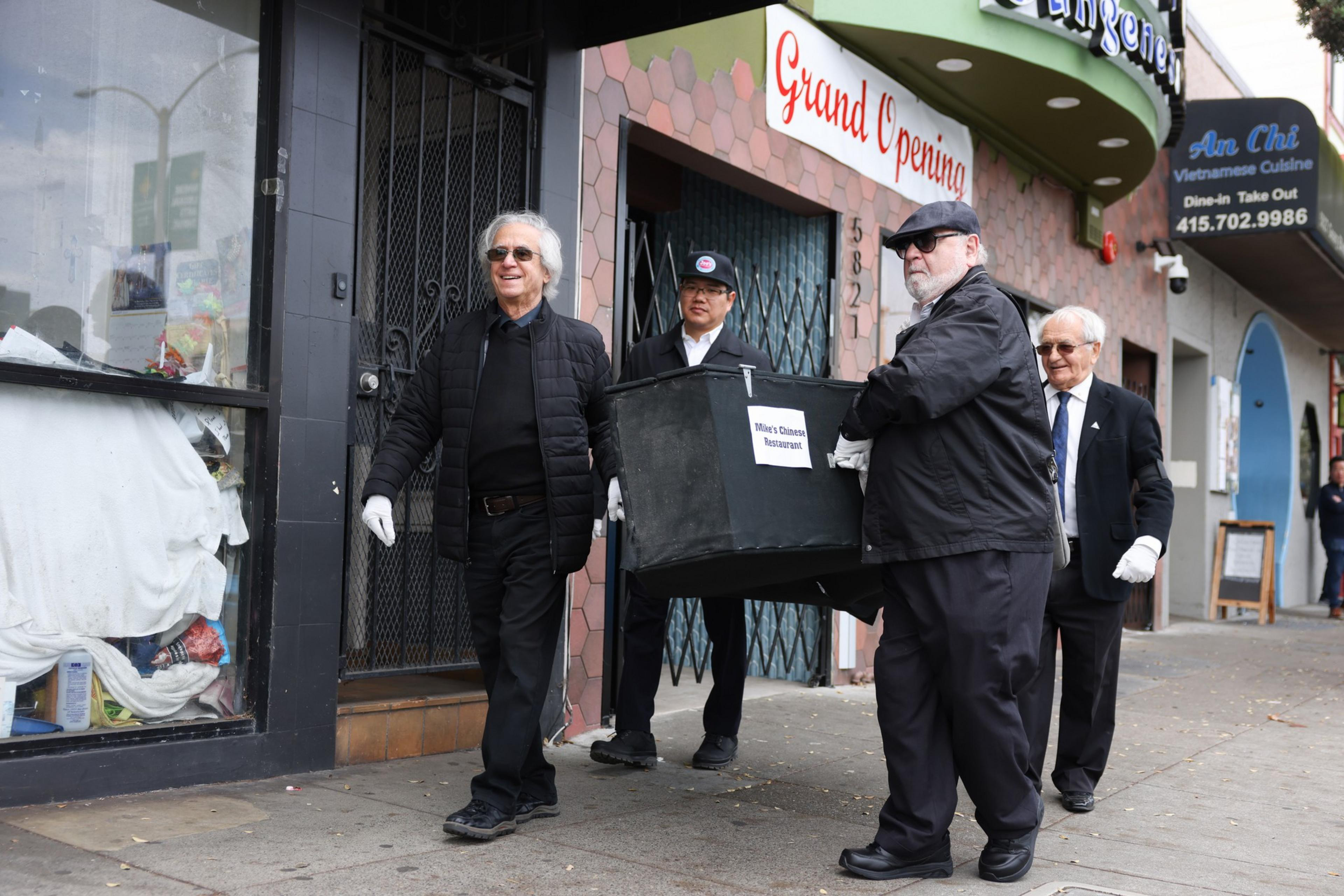 Four men in suits and white gloves carry a black box labeled "Mile's Cleanse Restaurant" on a city sidewalk near a storefront with a "Grand Opening" sign.