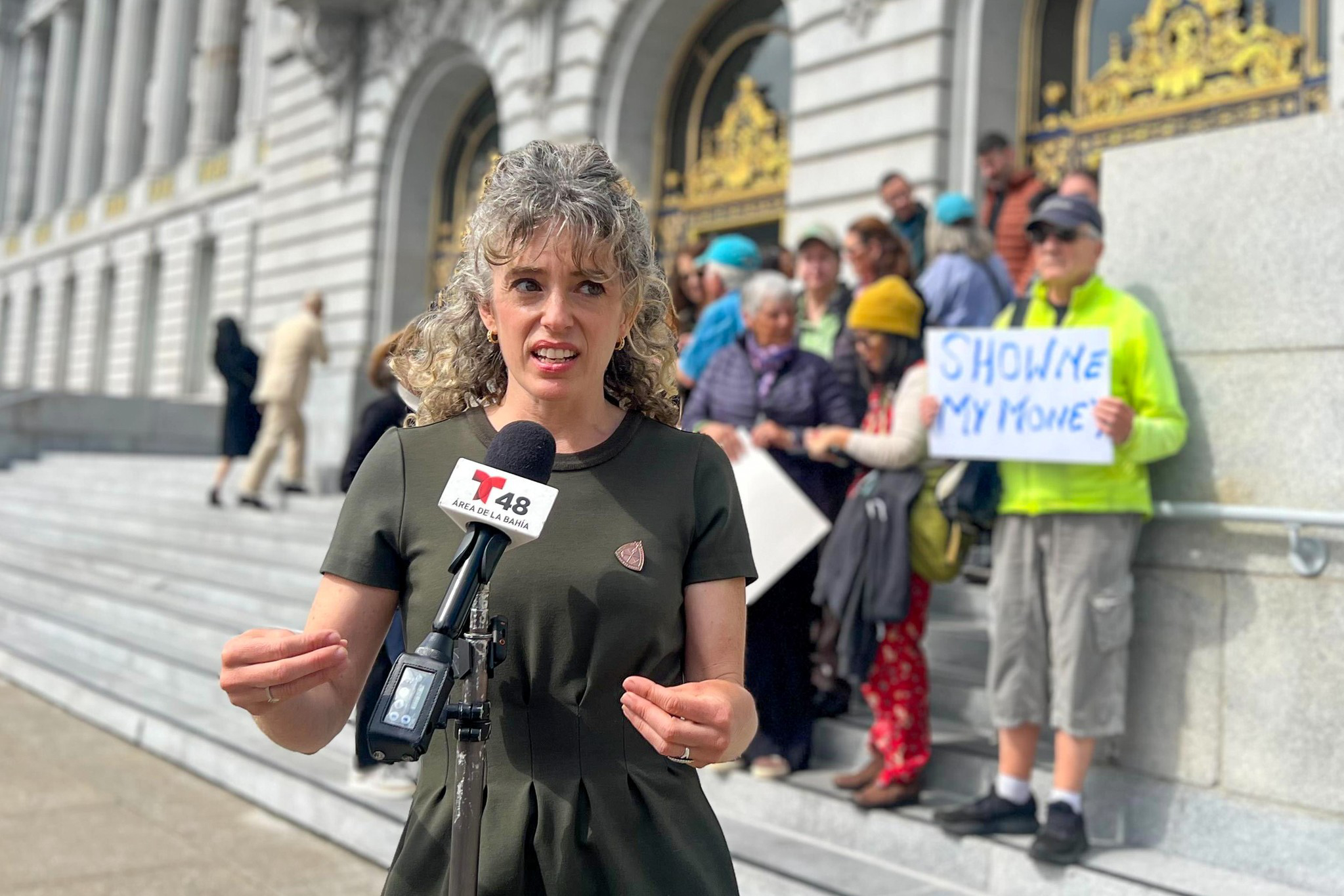 A woman stands in front of a microphone on the steps of a building. A group behind her holds a sign reading “Show me my money.” The building has ornate doors.