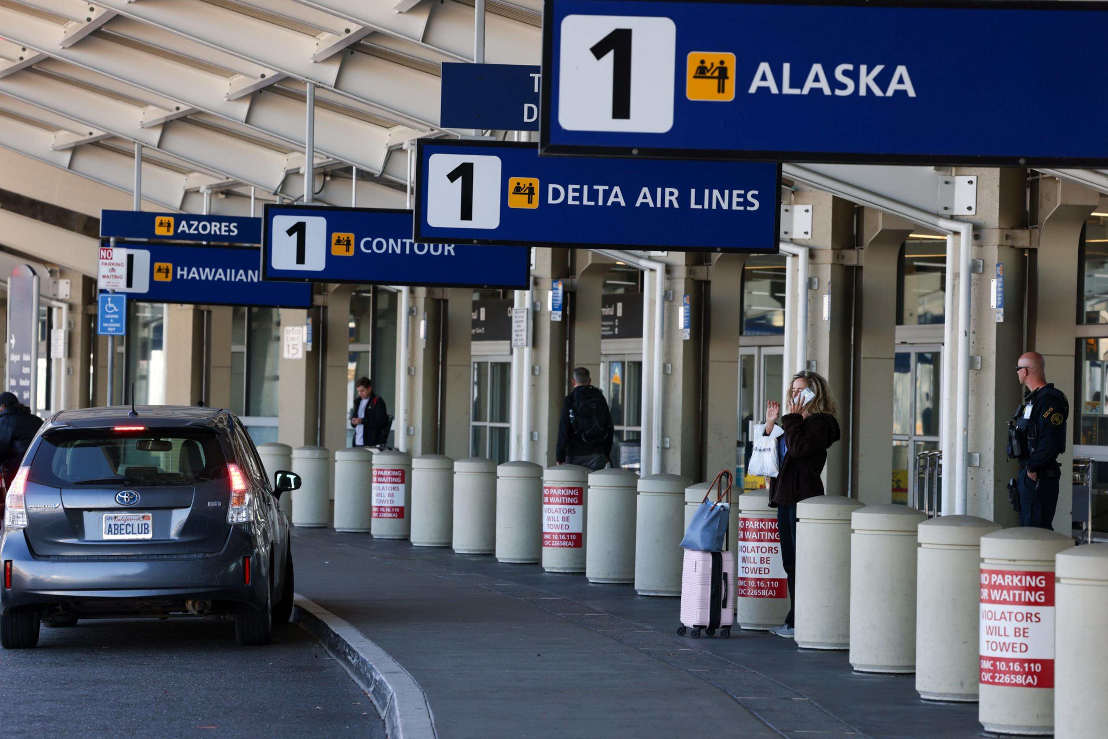 At an airport drop-off zone, people stand by signs for Alaska and Delta Air Lines; a car waits, and a security officer observes.