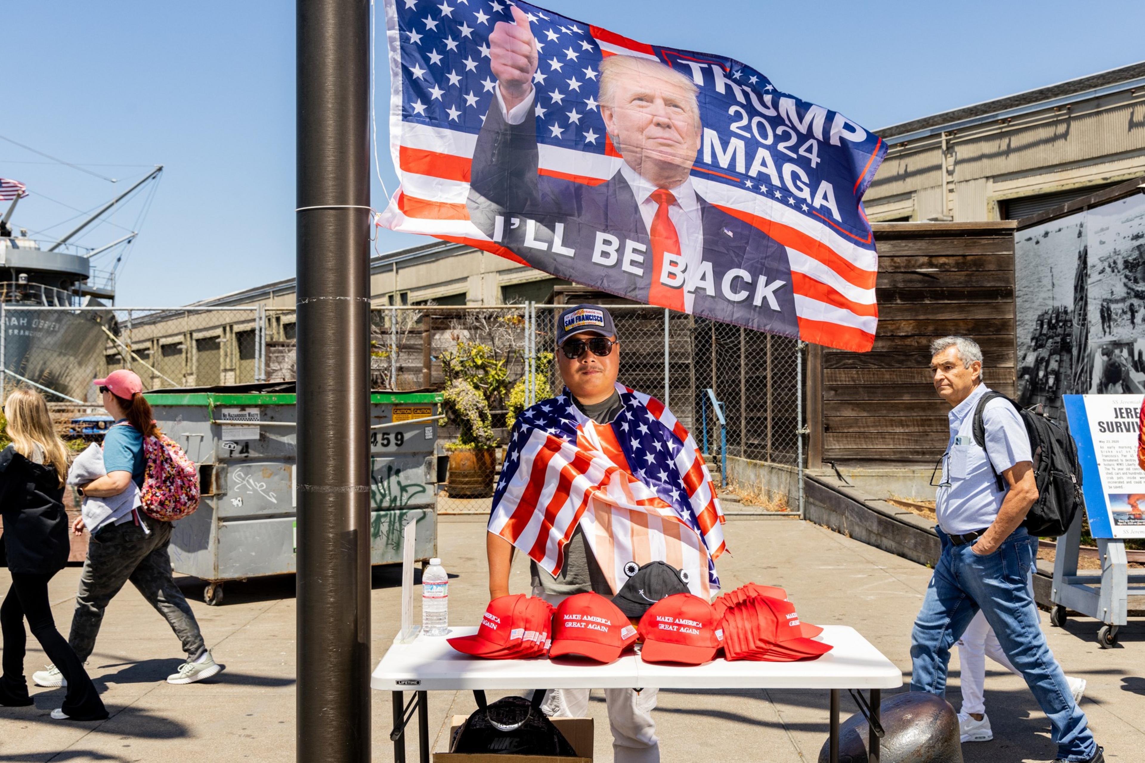 A man wearing an American flag stands by a table with red "Make America Great Again" hats, under a "Trump 2024" flag. People walk by in the background.