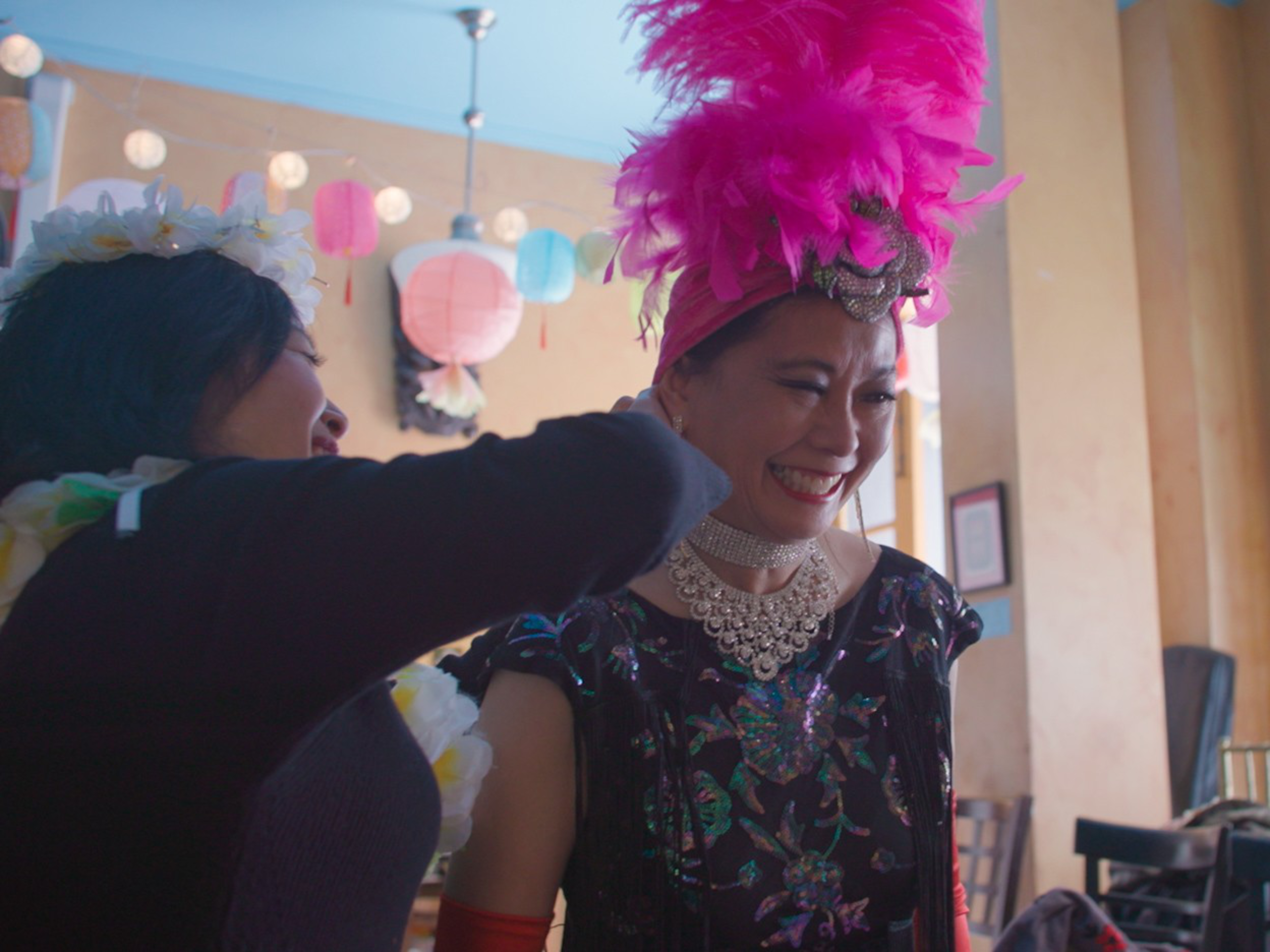 A smiling woman wearing a bright pink feathered headpiece and intricate necklace is being helped by another person in a flower crown.