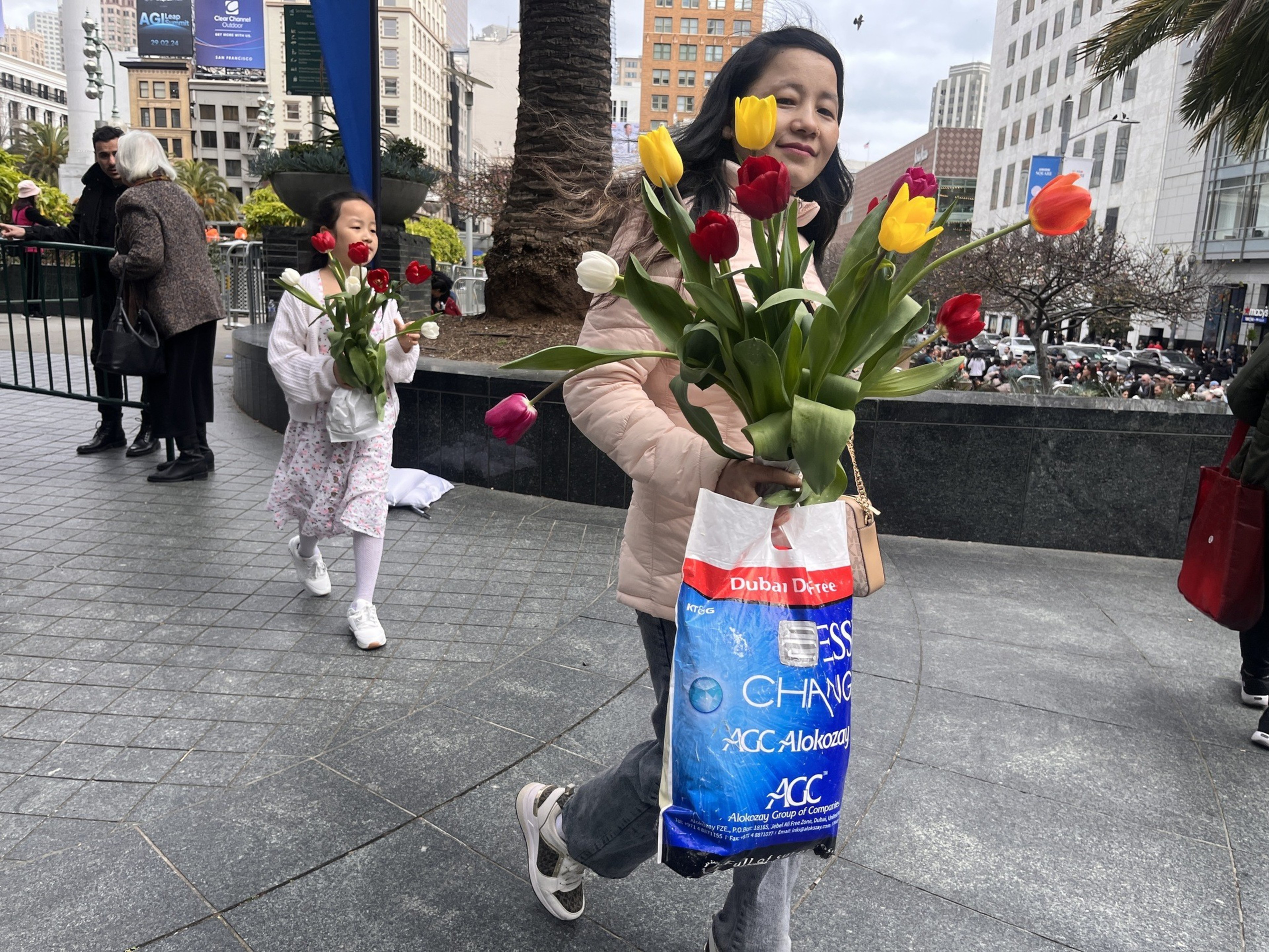 A woman holding colorful tulips in a plastic bag walks by a girl, both smiling, in an urban square.