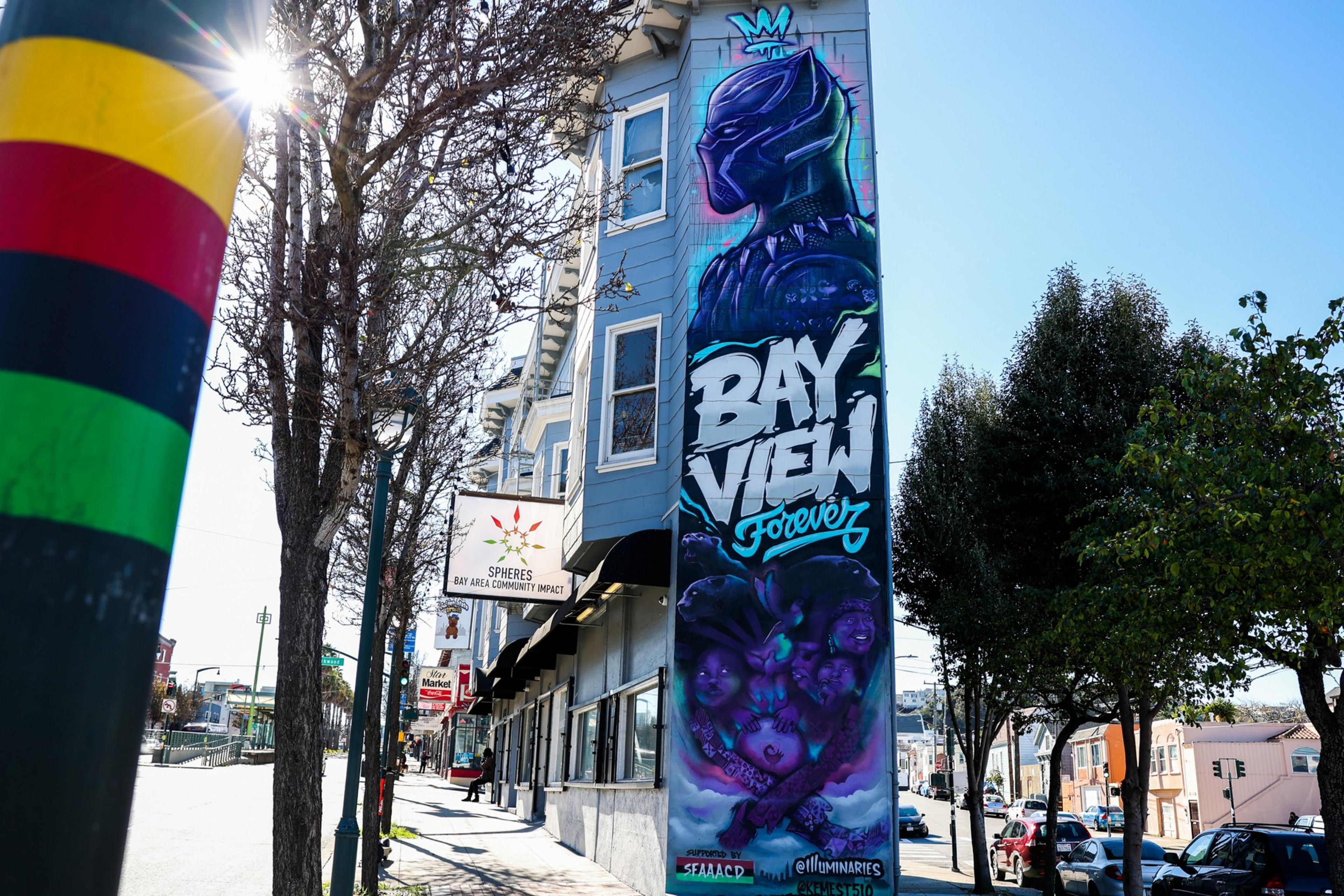 A mural with 'BAY VIEW Forever' on a building's side, trees, a clear sky, and a colorful pole on the left edge.