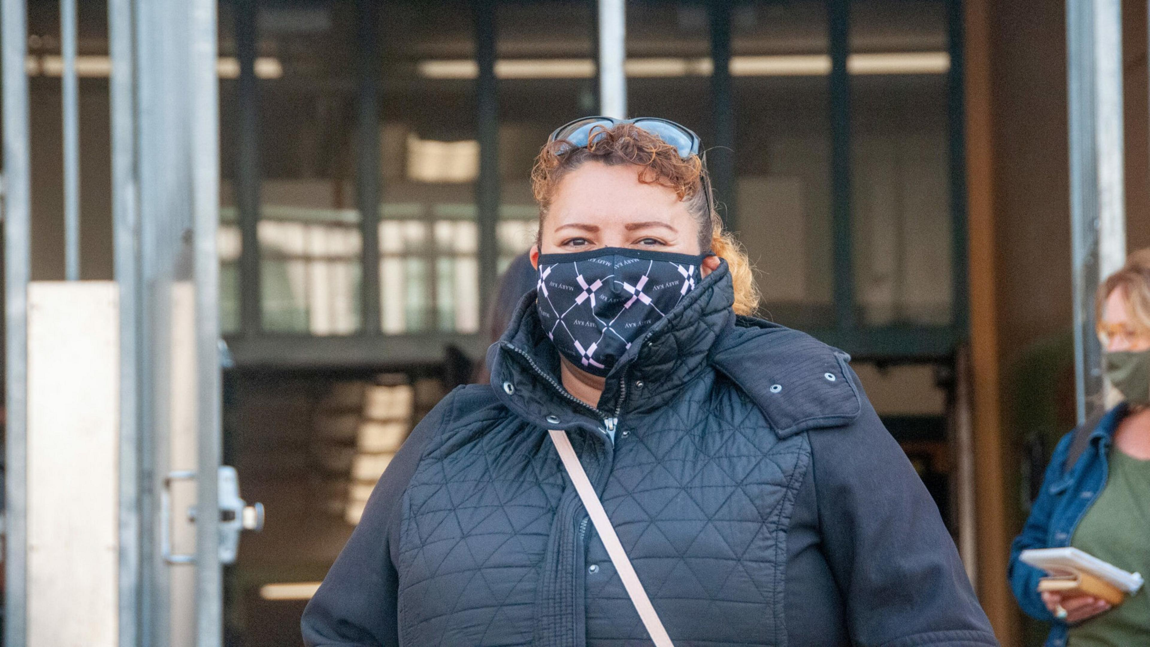 A woman wearing a black quilted jacket and patterned face mask stands outdoors with sunglasses on her head and a beige strap across her chest.