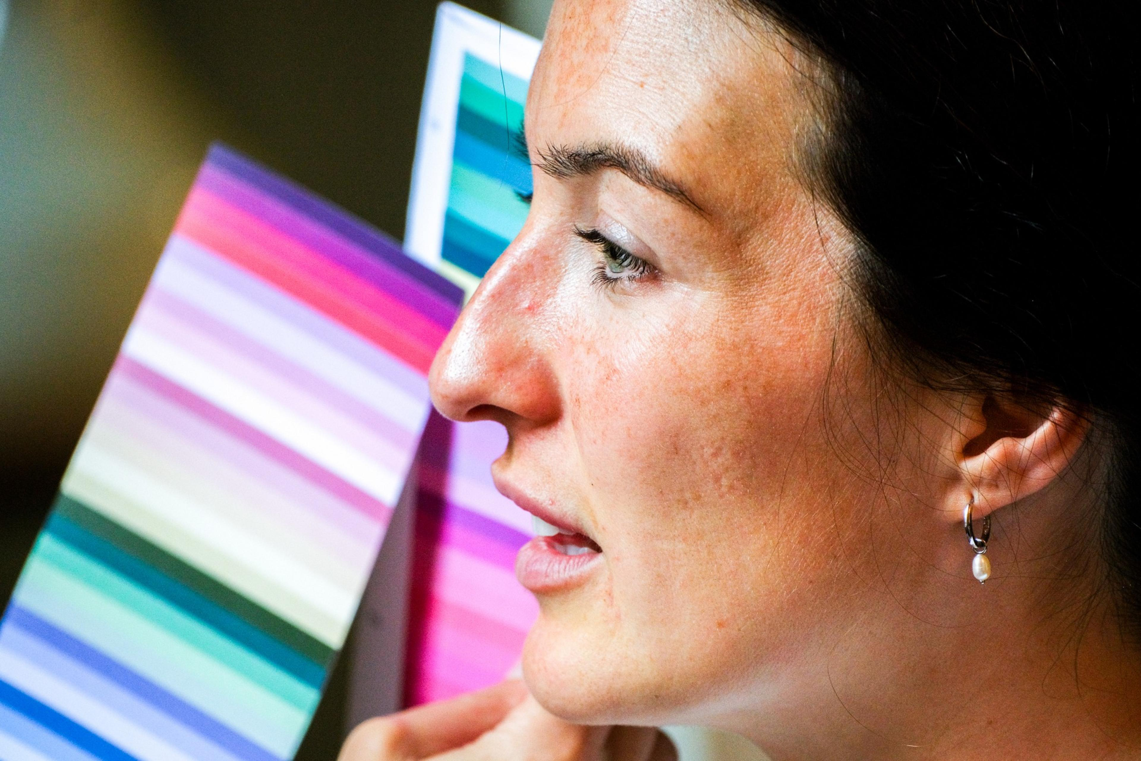 A woman with dark hair is seen in profile holding or using color swatches with various shades. She wears earrings and appears focused on the swatches.
